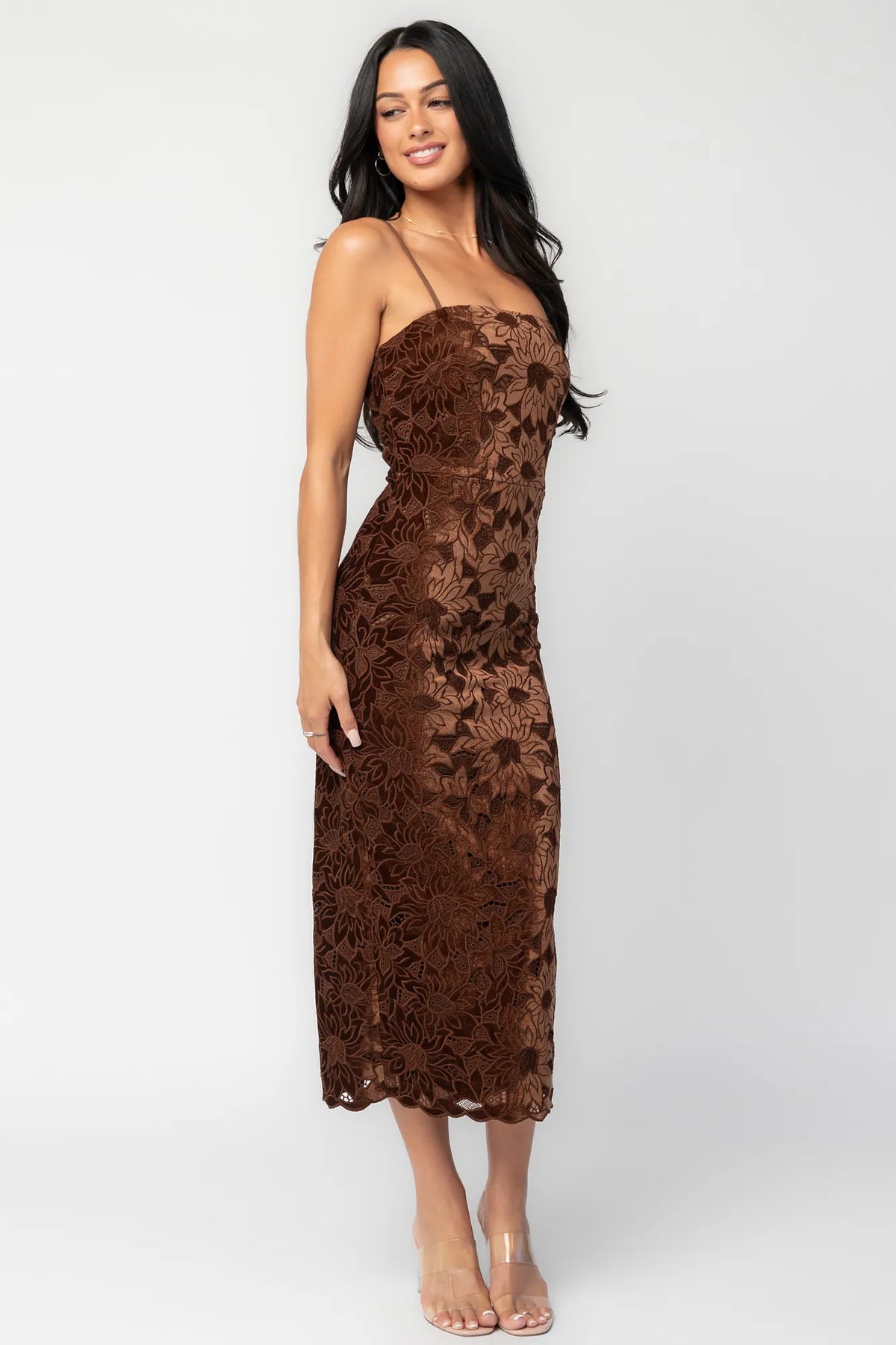 Model wearing a fitted espresso brown midi dress featuring an allover floral velvet lace overlay. The dress has a square neckline, adjustable spaghetti straps, and a body-skimming silhouette that falls below the knee with a scalloped hem.