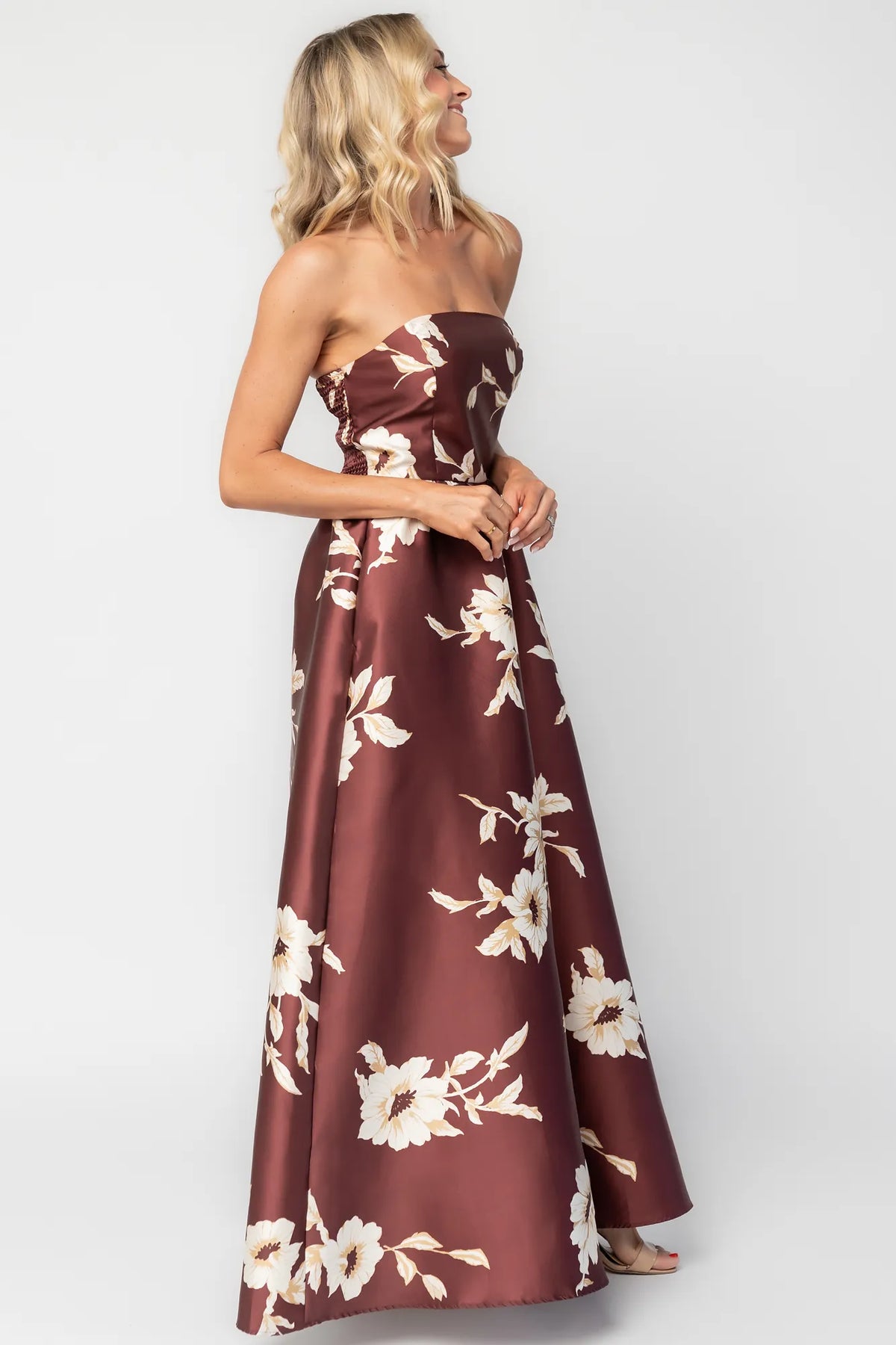 Model wearing a strapless maxi gown with a mahogany floral print and a fitted bodice. A rosette detail accents the neckline, while the flowing floor-length skirt creates elegant movement.