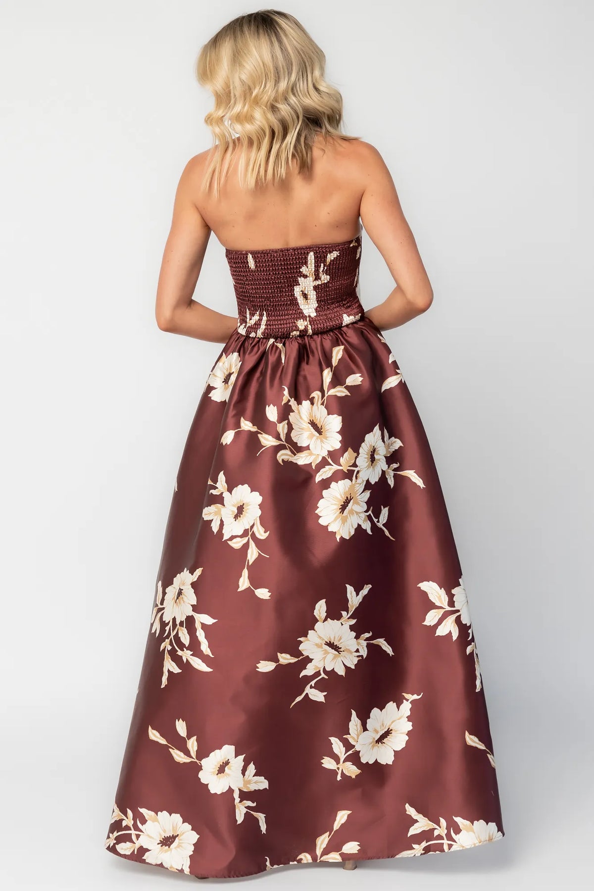 Model wearing a strapless maxi gown with a mahogany floral print and a fitted bodice. A rosette detail accents the neckline, while the flowing floor-length skirt creates elegant movement.