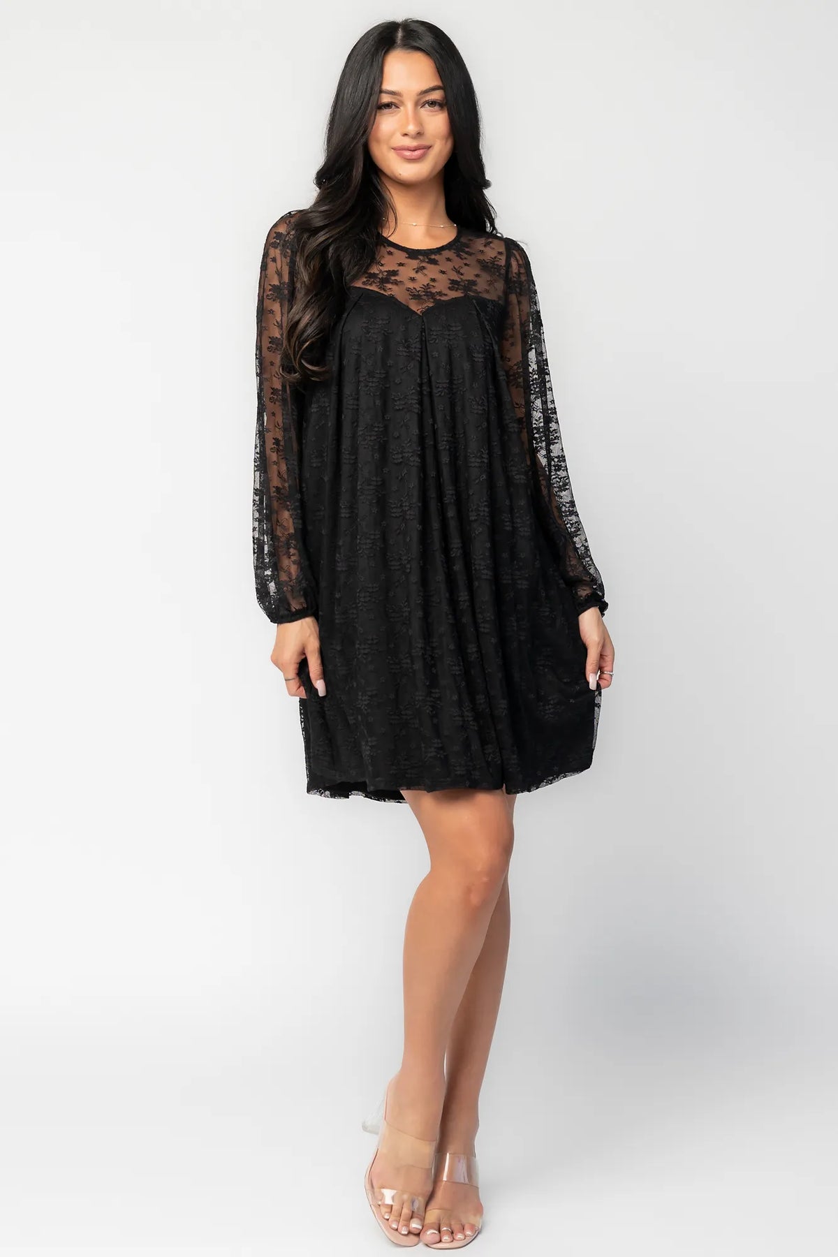 Model wearing a black short lace dress with a sheer floral lace yoke and long sheer sleeves. The dress features a sweetheart-style lined bodice beneath the lace overlay, a round neckline, and a flowy silhouette that falls above the knee.