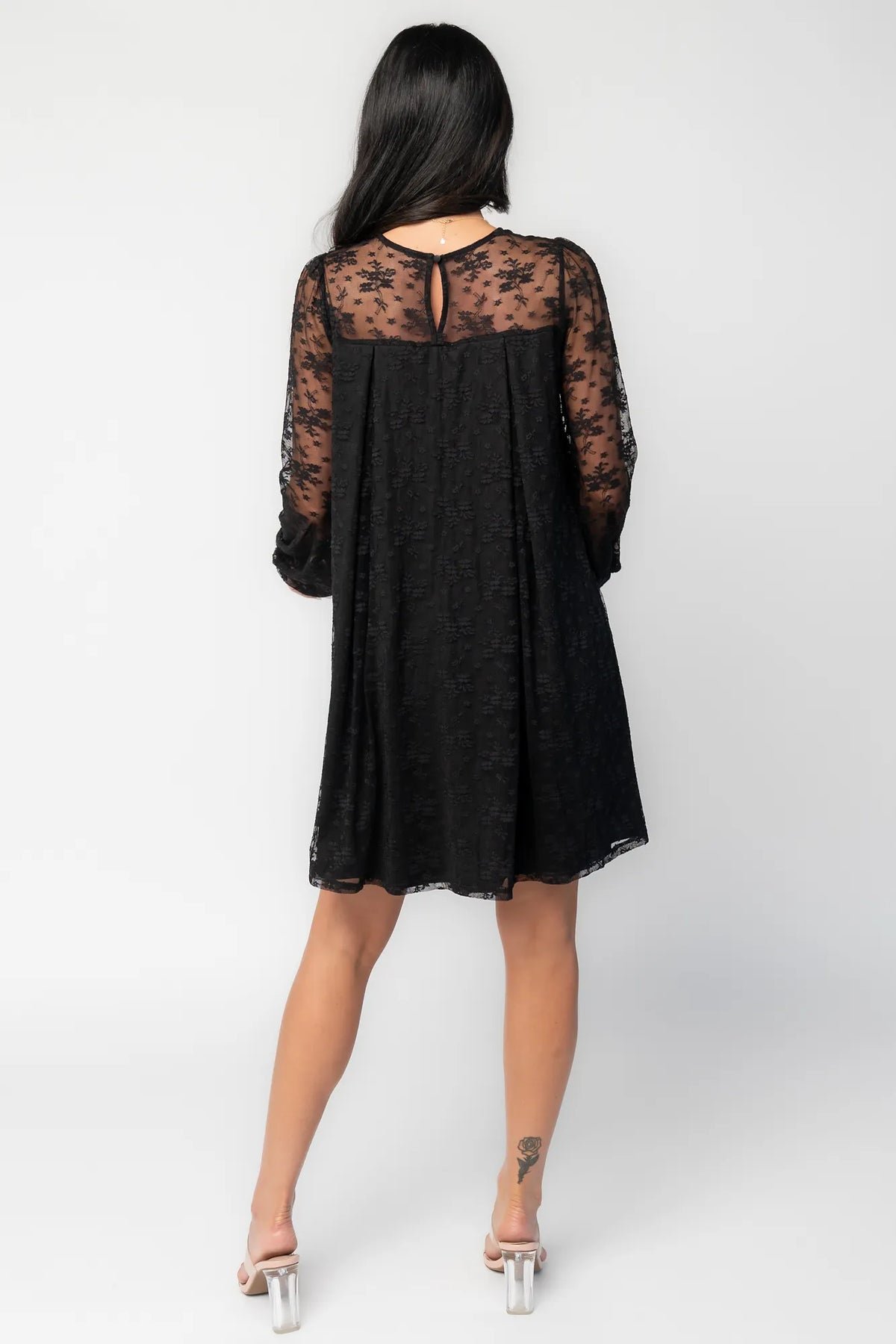 Model wearing a black short lace dress with a sheer floral lace yoke and long sheer sleeves. The dress features a sweetheart-style lined bodice beneath the lace overlay, a round neckline, and a flowy silhouette that falls above the knee.