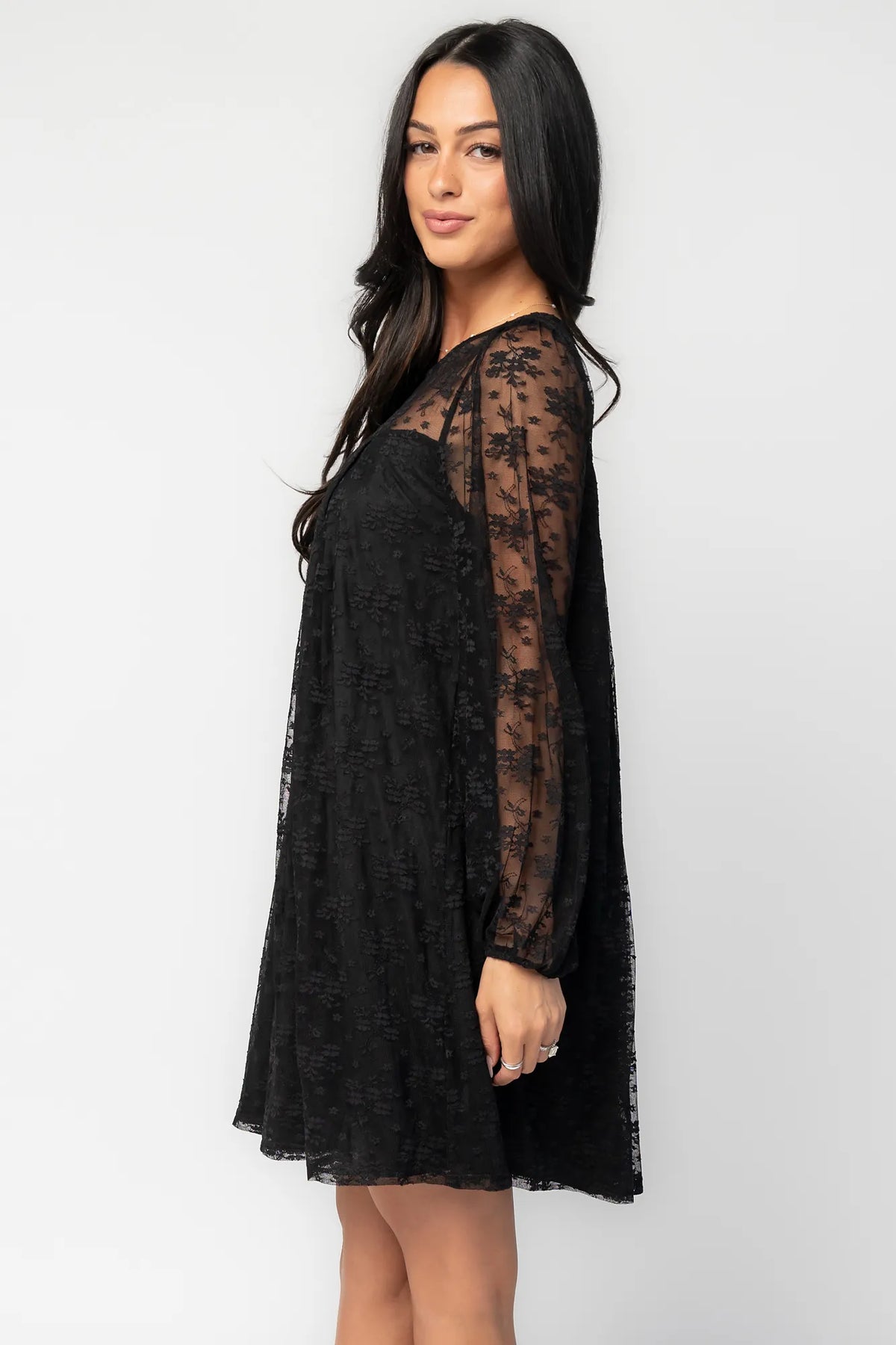 Model wearing a black short lace dress with a sheer floral lace yoke and long sheer sleeves. The dress features a sweetheart-style lined bodice beneath the lace overlay, a round neckline, and a flowy silhouette that falls above the knee.