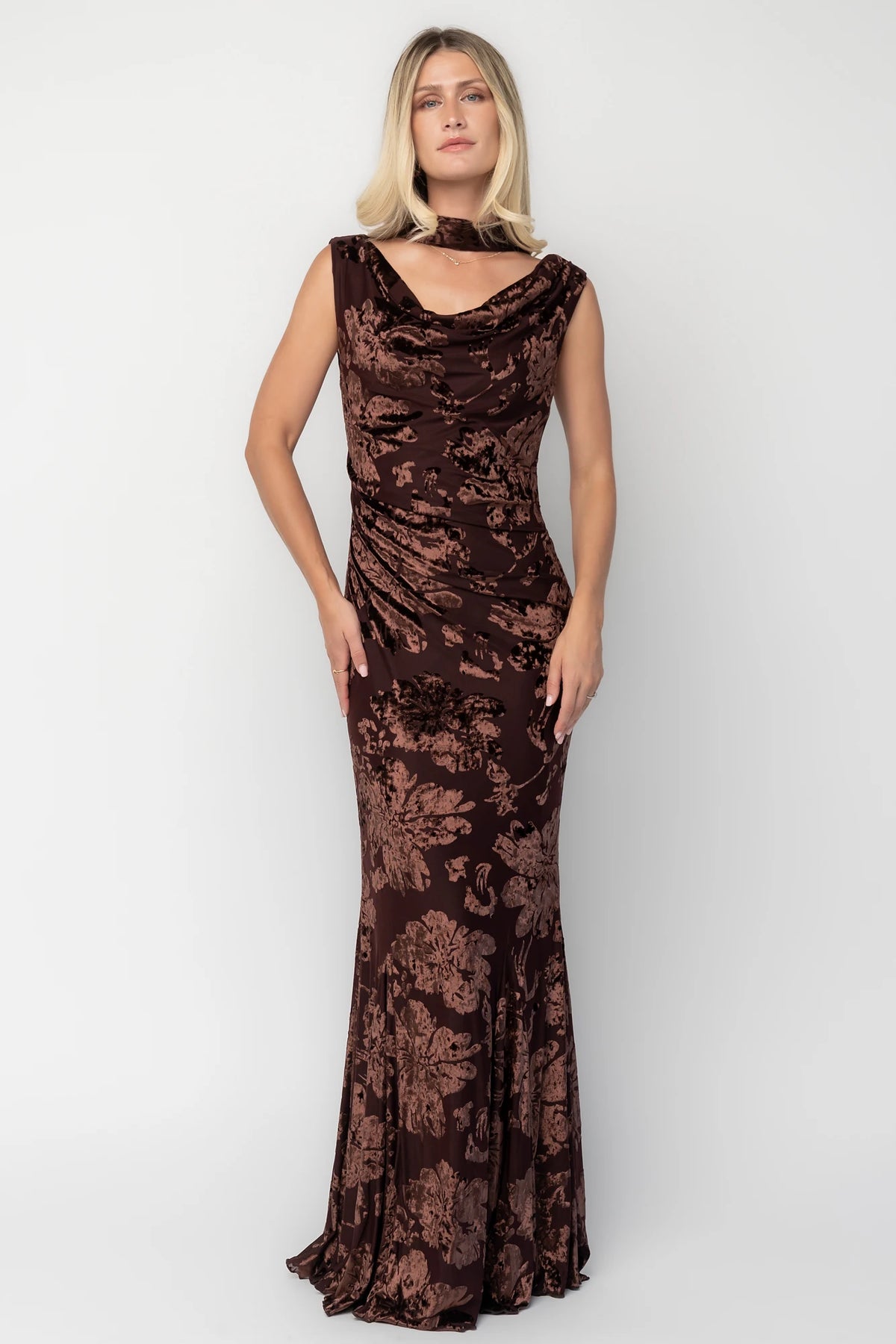 Model wearing a brown velvet burnout floral maxi dress with a draped cowl neckline, scarf detail, side ruching, and a flowing mermaid silhouette.