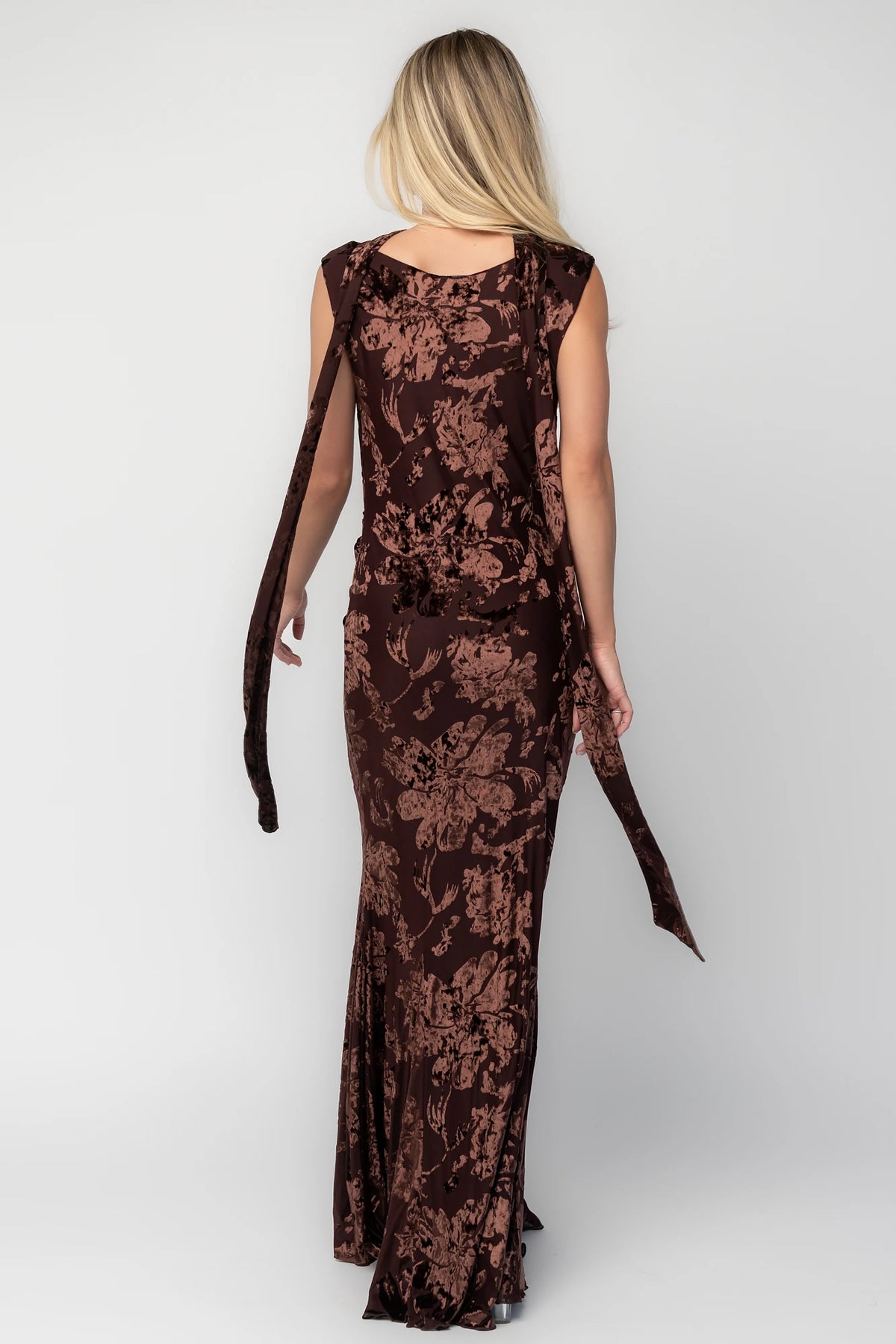 Model wearing a brown velvet burnout floral maxi dress with a draped cowl neckline, scarf detail, side ruching, and a flowing mermaid silhouette.