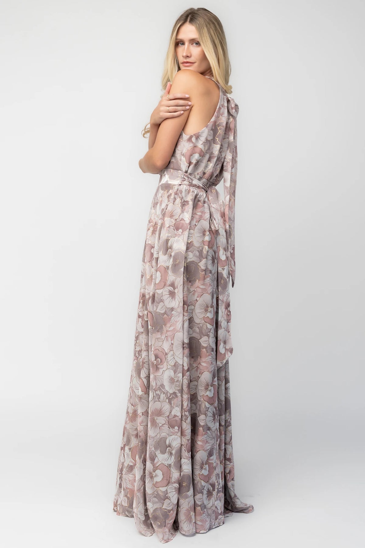 Model wearing the Tinsley Metallic Maxi Dress in Dusty Mauve featuring a high pleated halter neckline, defined waistband, tie-back keyhole detail, and a flowing chiffon maxi skirt with a dusty mauve floral print and subtle metallic shimmer.