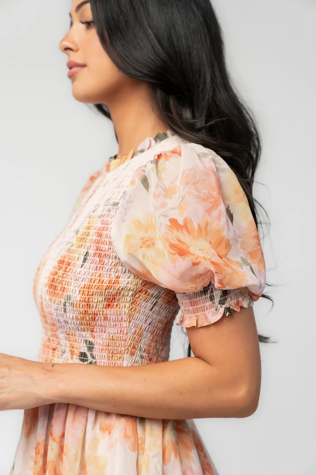 Model wearing an ivory floral short dress with a fully smocked bodice, ruffled round neckline, and short puff sleeves.
