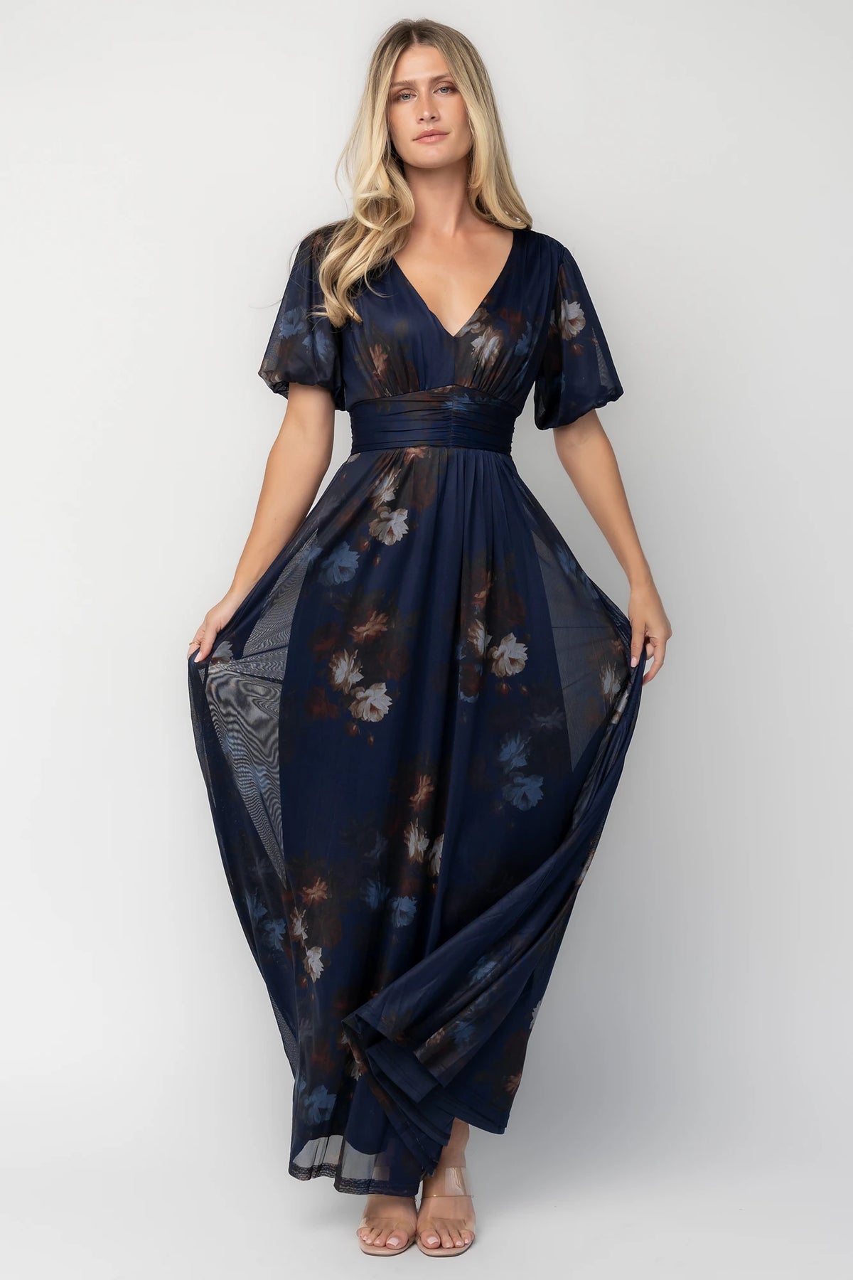 Model wearing the Whitney Mesh Maxi Dress in Navy, a navy floral mesh maxi dress with a deep V-neckline, short flutter sleeves, an empire waist, and a flowing A-line skirt with side pockets.