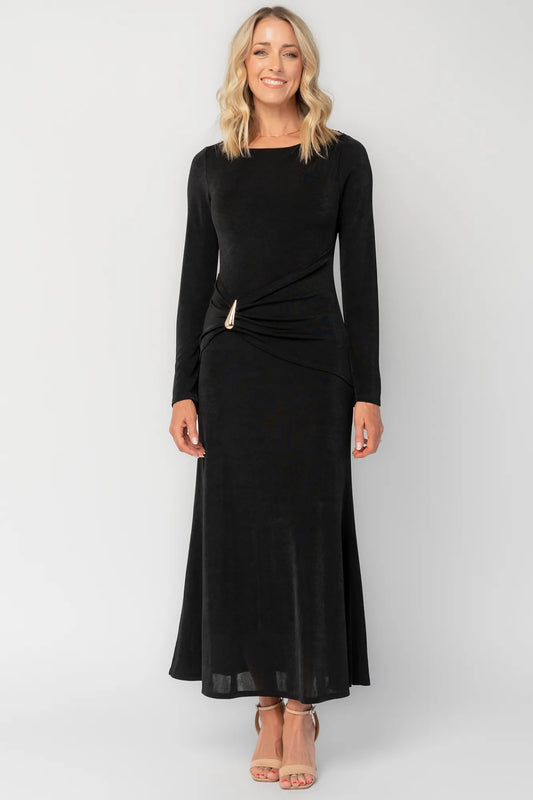 Model wearing a black long-sleeve maxi dress with a fitted silhouette and round neckline. Draped fabric gathers at the waist and is accented by a gold-tone metal detail.