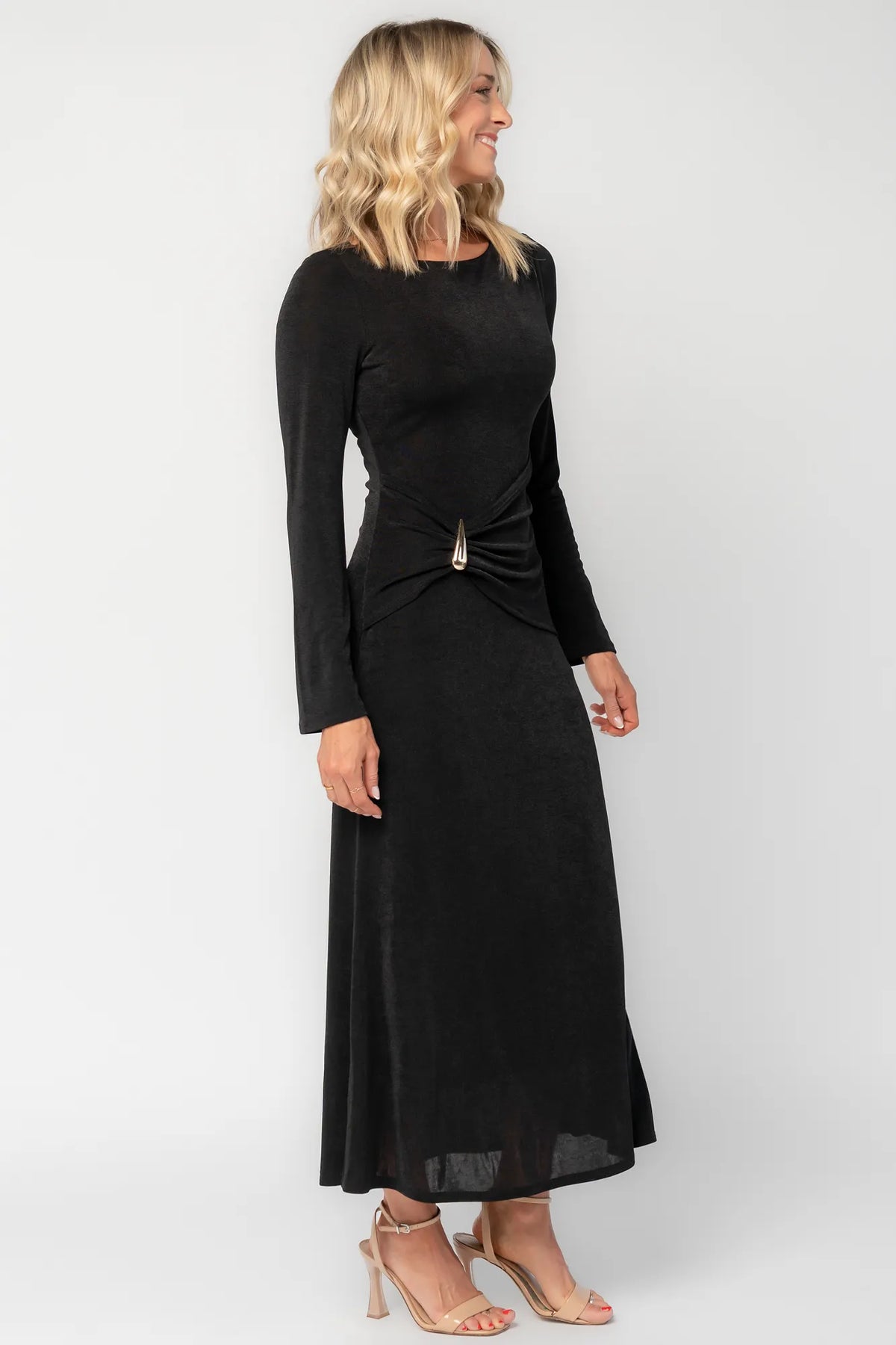 Model wearing a black long-sleeve maxi dress with a fitted silhouette and round neckline. Draped fabric gathers at the waist and is accented by a gold-tone metal detail.