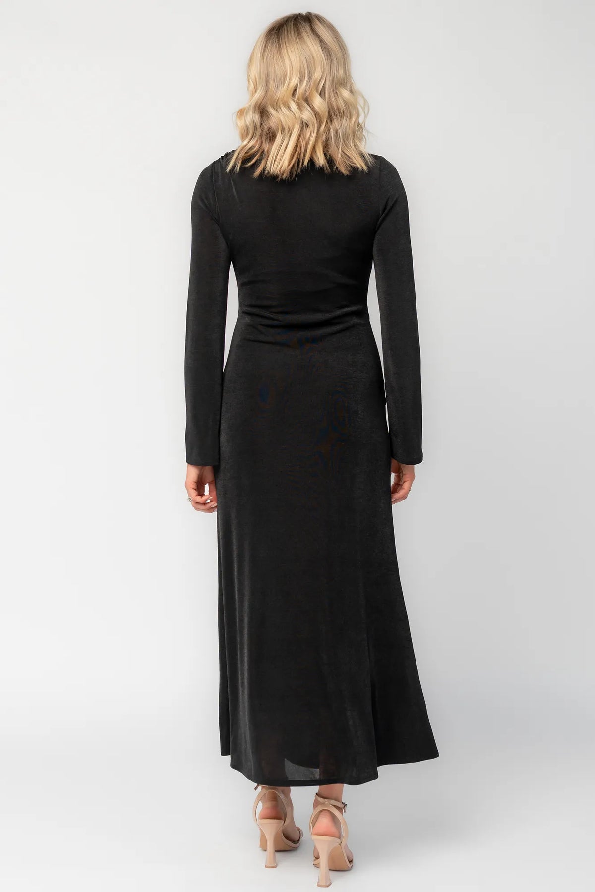 Model wearing a black long-sleeve maxi dress with a fitted silhouette and round neckline. Draped fabric gathers at the waist and is accented by a gold-tone metal detail.