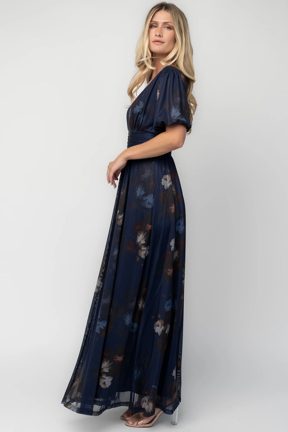 Model wearing the Whitney Mesh Maxi Dress in Navy, a navy floral mesh maxi dress with a deep V-neckline, short flutter sleeves, an empire waist, and a flowing A-line skirt with side pockets.