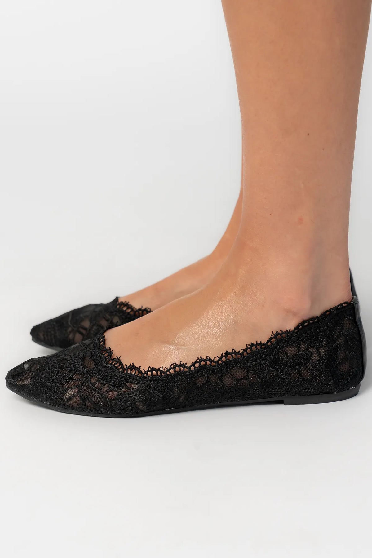 Model wearing black lace ballet flats with a pointed toe, sheer lace construction, and scalloped edges. The feminine flat silhouette offers an elegant, lightweight look.