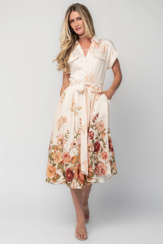 Model wearing a cream floral midi dress with a collared neckline, functional button-front bodice, short sleeves, a removable waist tie, and a flowing midi-length skirt.