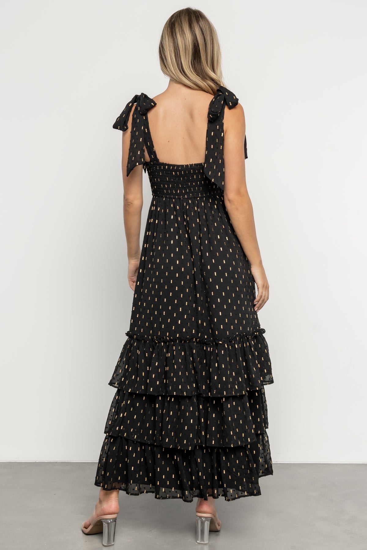 Model in Kassia Maxi Dress showing ruffle tiers, empire waist silhouette, and gold accents on black fabric.