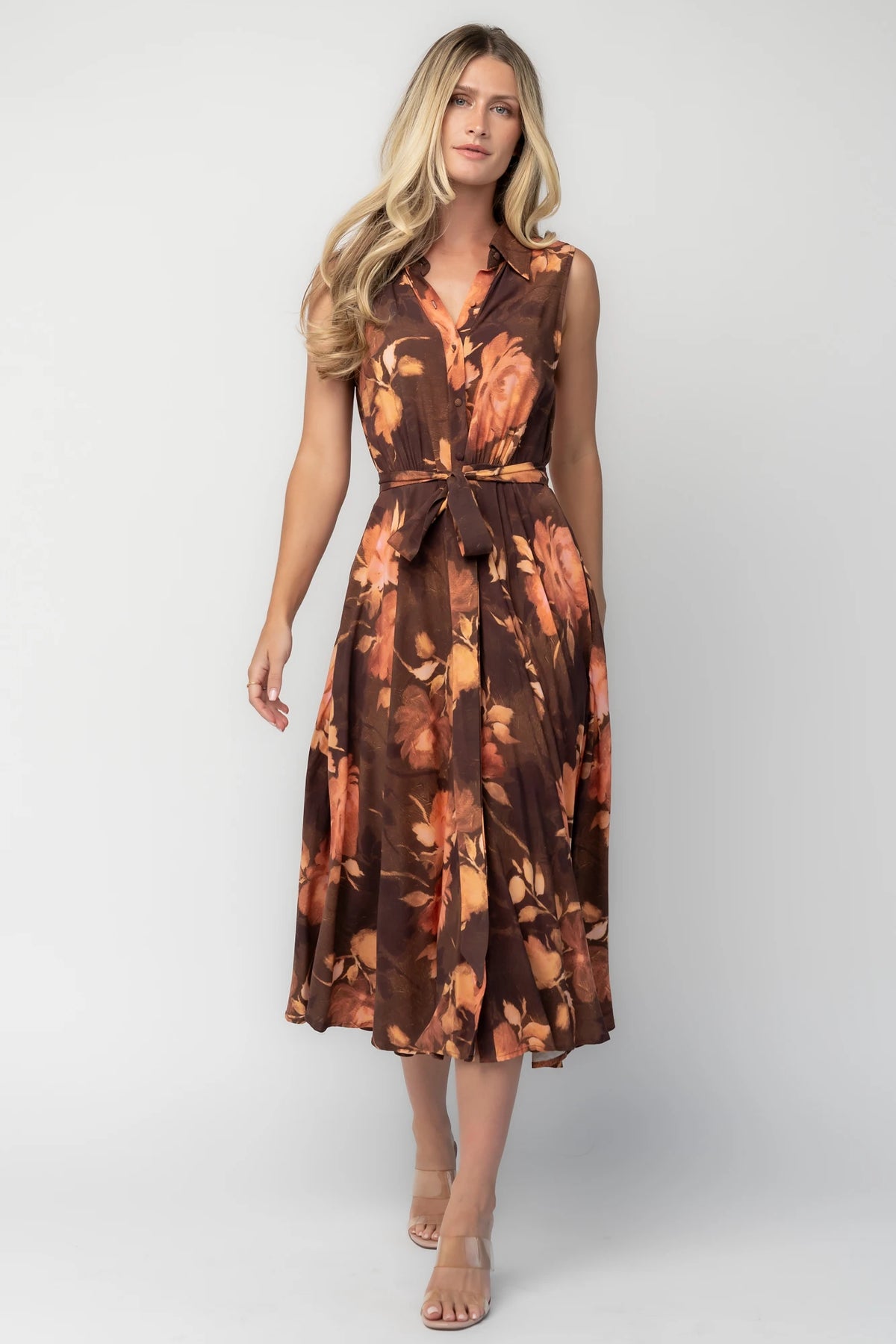 Model wearing the Tess Midi Dress in Brown + Rust featuring a collared neckline, functional button-front bodice, sleeveless design, removable waist tie, elastic back waist, hidden side pockets, and a flowing midi skirt with a brown background and warm rust floral print.