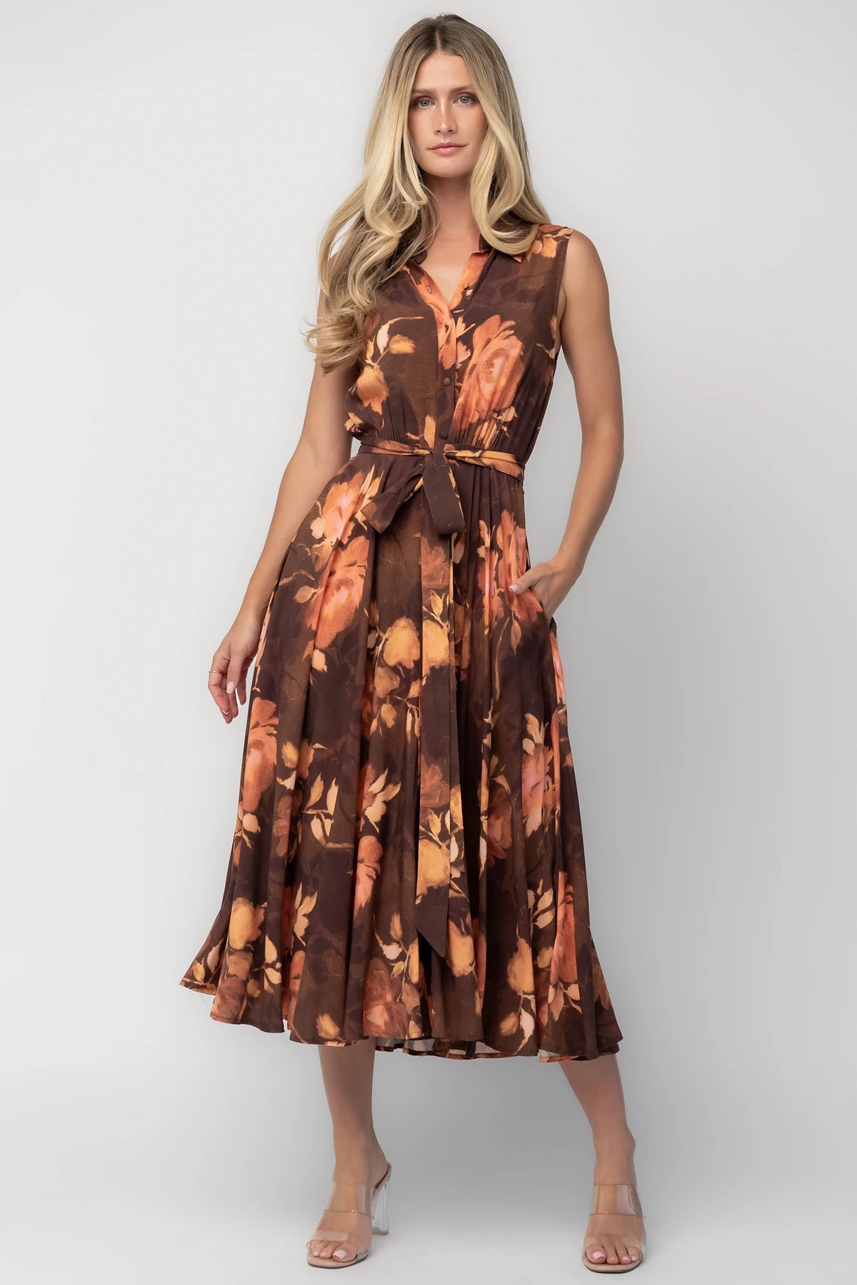 Model wearing the Tess Midi Dress in Brown + Rust featuring a collared neckline, functional button-front bodice, sleeveless design, removable waist tie, elastic back waist, hidden side pockets, and a flowing midi skirt with a brown background and warm rust floral print.