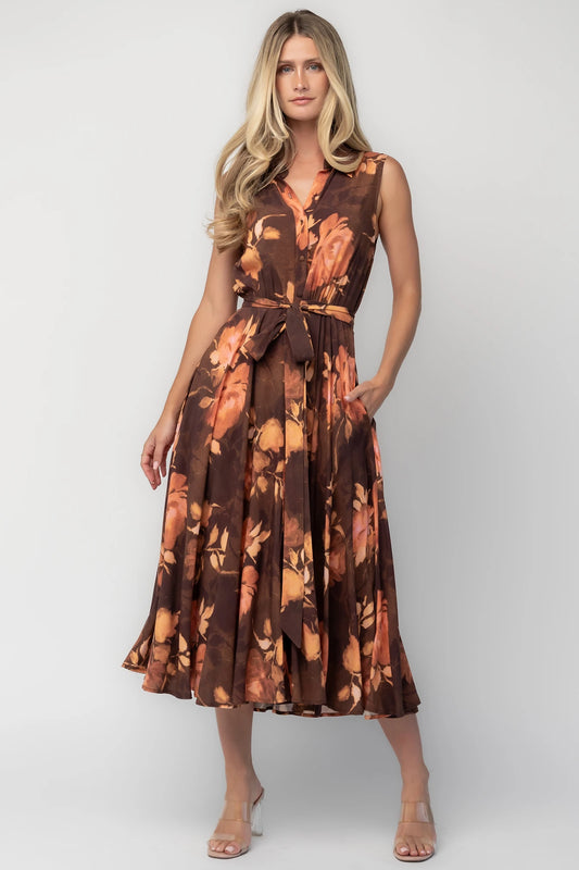 Model wearing the Tess Midi Dress in Brown + Rust featuring a collared neckline, functional button-front bodice, sleeveless design, removable waist tie, elastic back waist, hidden side pockets, and a flowing midi skirt with a brown background and warm rust floral print.