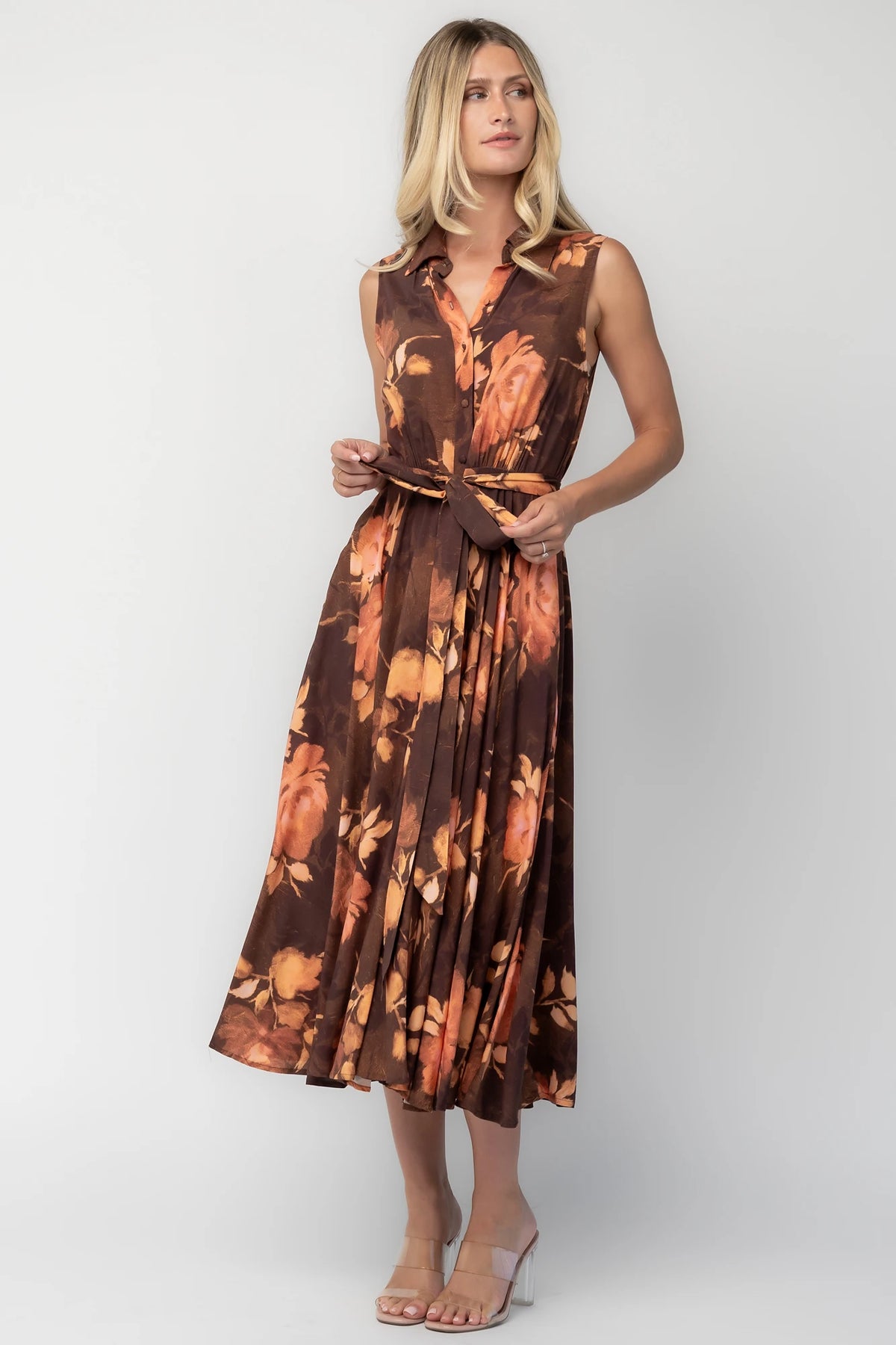 Model wearing the Tess Midi Dress in Brown + Rust featuring a collared neckline, functional button-front bodice, sleeveless design, removable waist tie, elastic back waist, hidden side pockets, and a flowing midi skirt with a brown background and warm rust floral print.