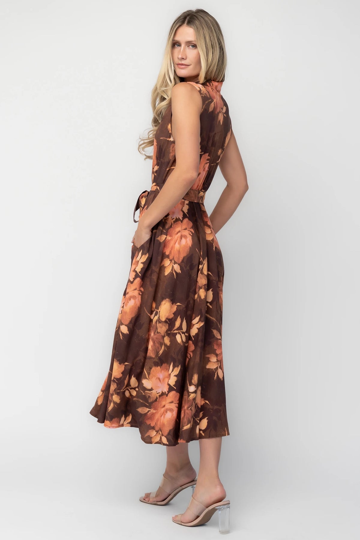 Model wearing the Tess Midi Dress in Brown + Rust featuring a collared neckline, functional button-front bodice, sleeveless design, removable waist tie, elastic back waist, hidden side pockets, and a flowing midi skirt with a brown background and warm rust floral print.