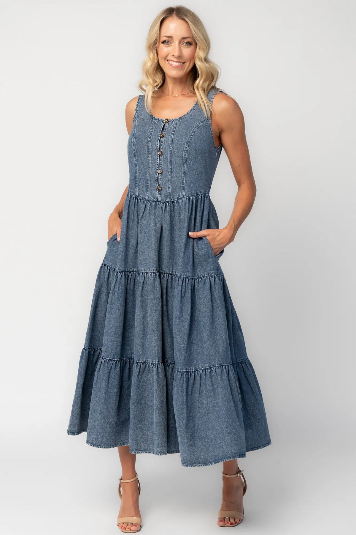 Woman wearing a sleeveless blue denim midi dress with a round neckline, button-front bodice, side pockets, and a tiered skirt.
