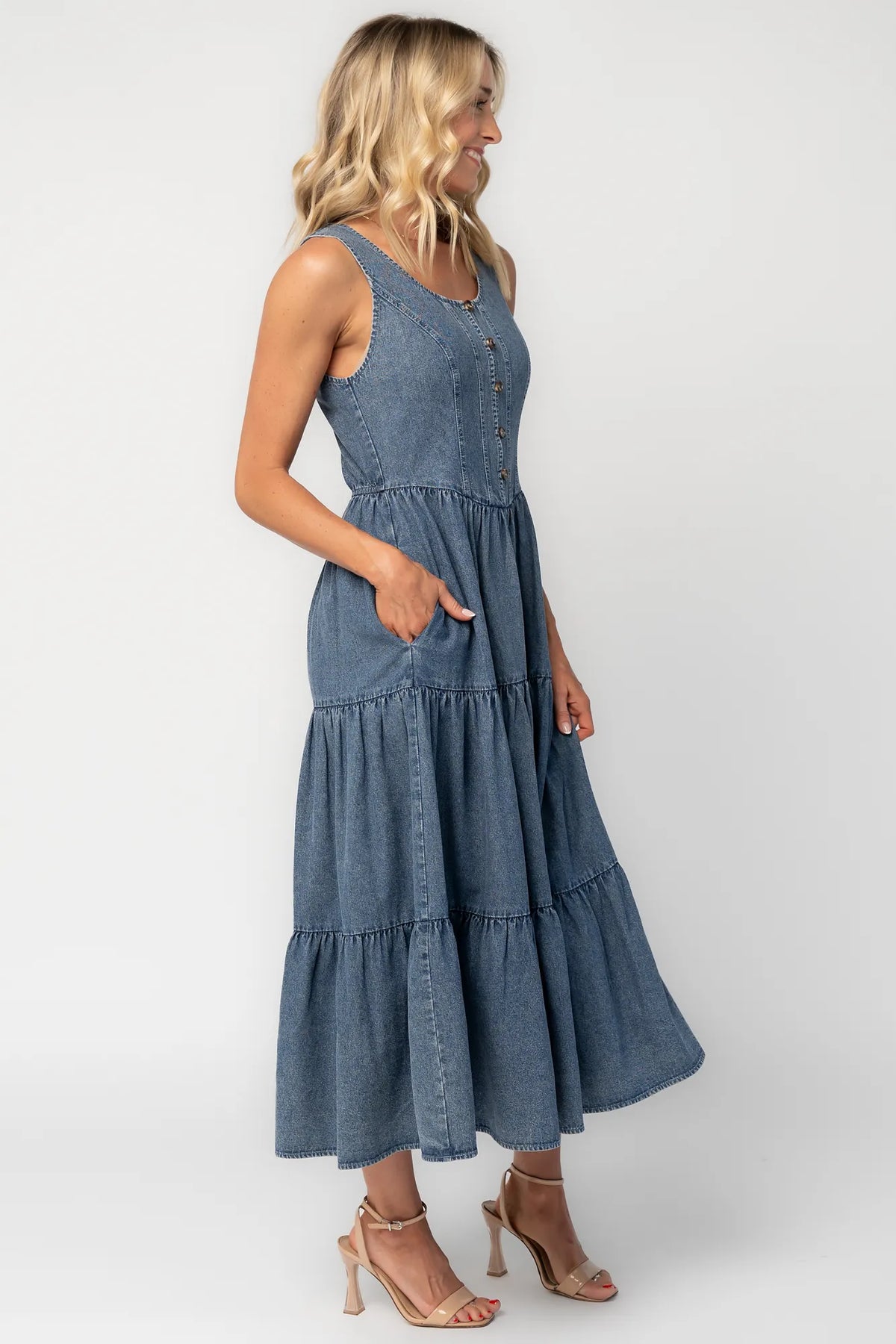 Side image of a woman wearing a sleeveless blue denim midi dress with a round neckline, button-front bodice, side pockets, and a tiered skirt.