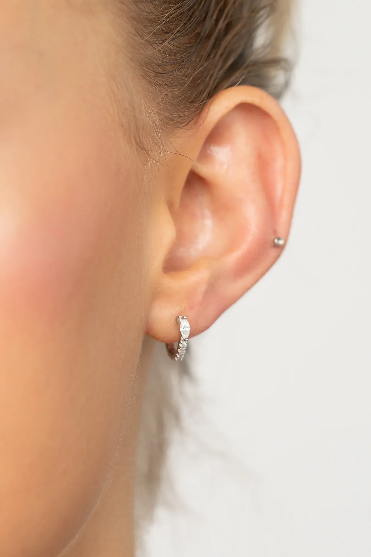 Model wearing petite sterling silver huggie hoop earrings featuring a marquise-cut crystal accent and a row of sparkling stones along the front. 