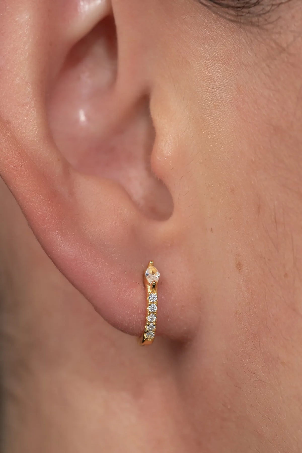 Model wearing petite gold huggie hoop earrings featuring a marquise-cut crystal accent and a row of sparkling stones along the front. 