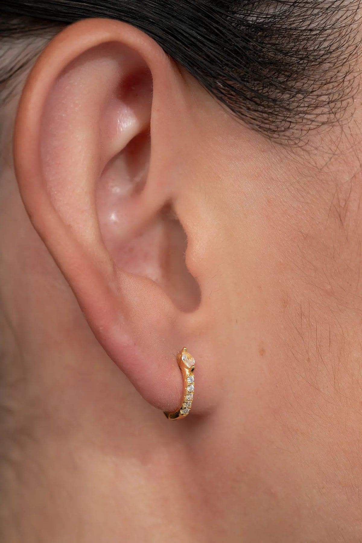 Model wearing petite gold huggie hoop earrings featuring a marquise-cut crystal accent and a row of sparkling stones along the front. 