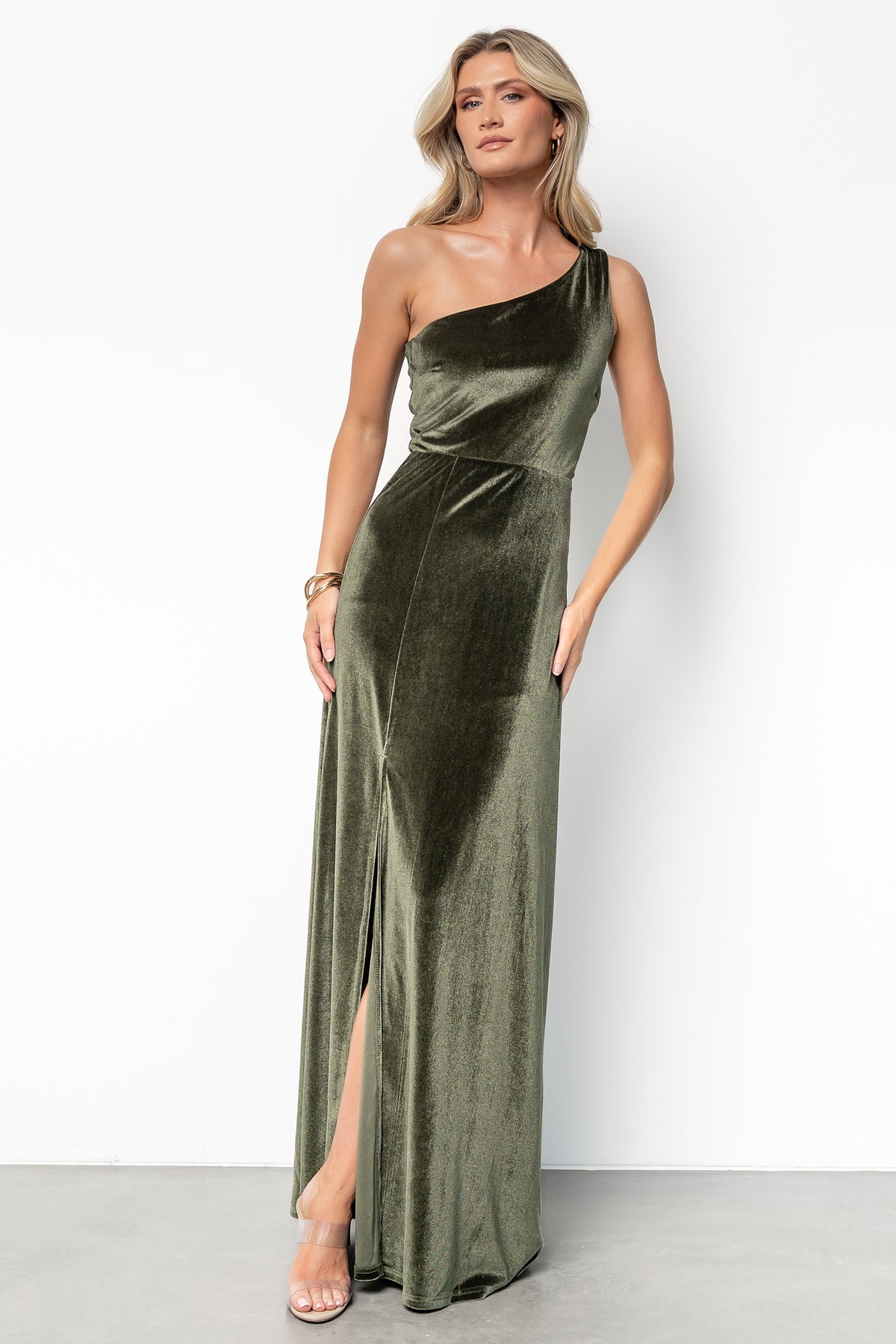 Full-length image showing olive velvet one-shoulder gown from a slight angle; asymmetrical neckline and slit to mid-thigh emphasize movement and drape.