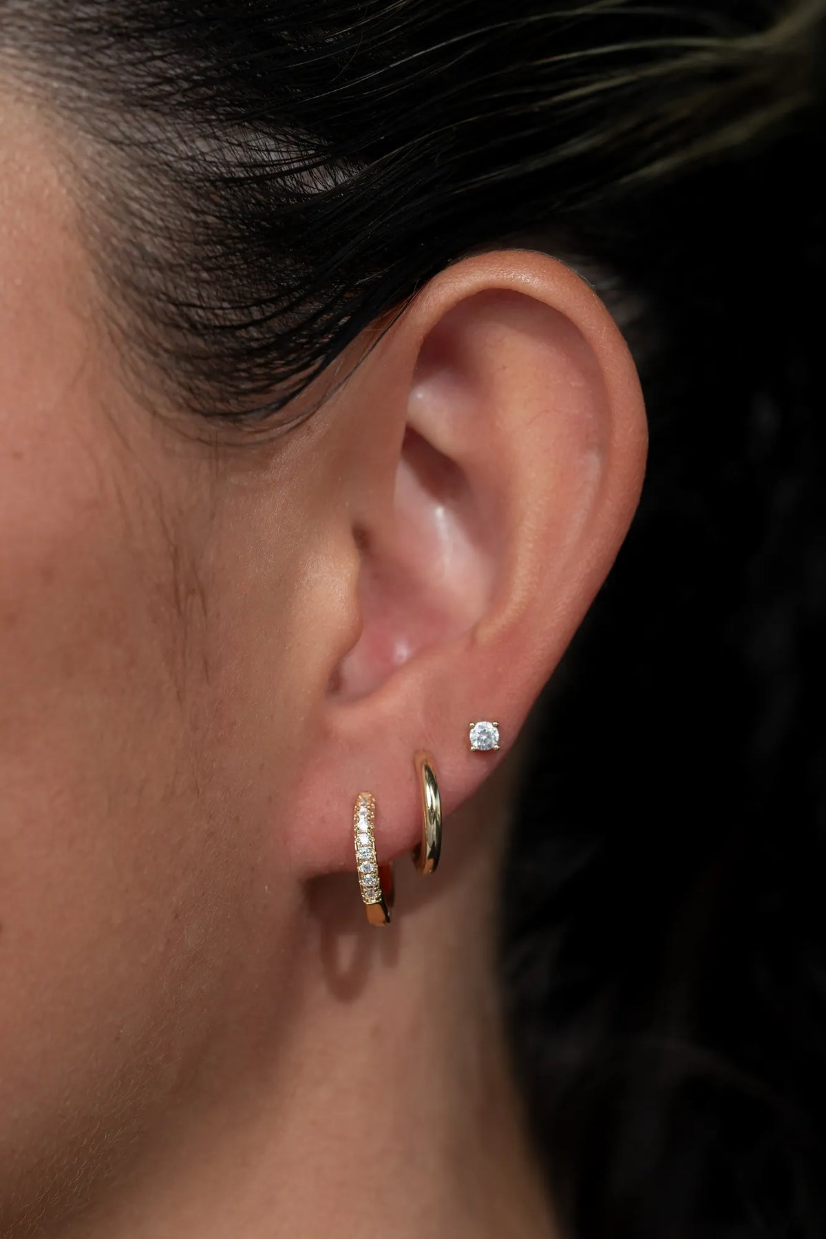 Model wearing a coordinated earring stack featuring gold-tone huggie hoop earrings with sparkling crystal accents, polished gold-tone huggie hoops, and round crystal stud earrings. 