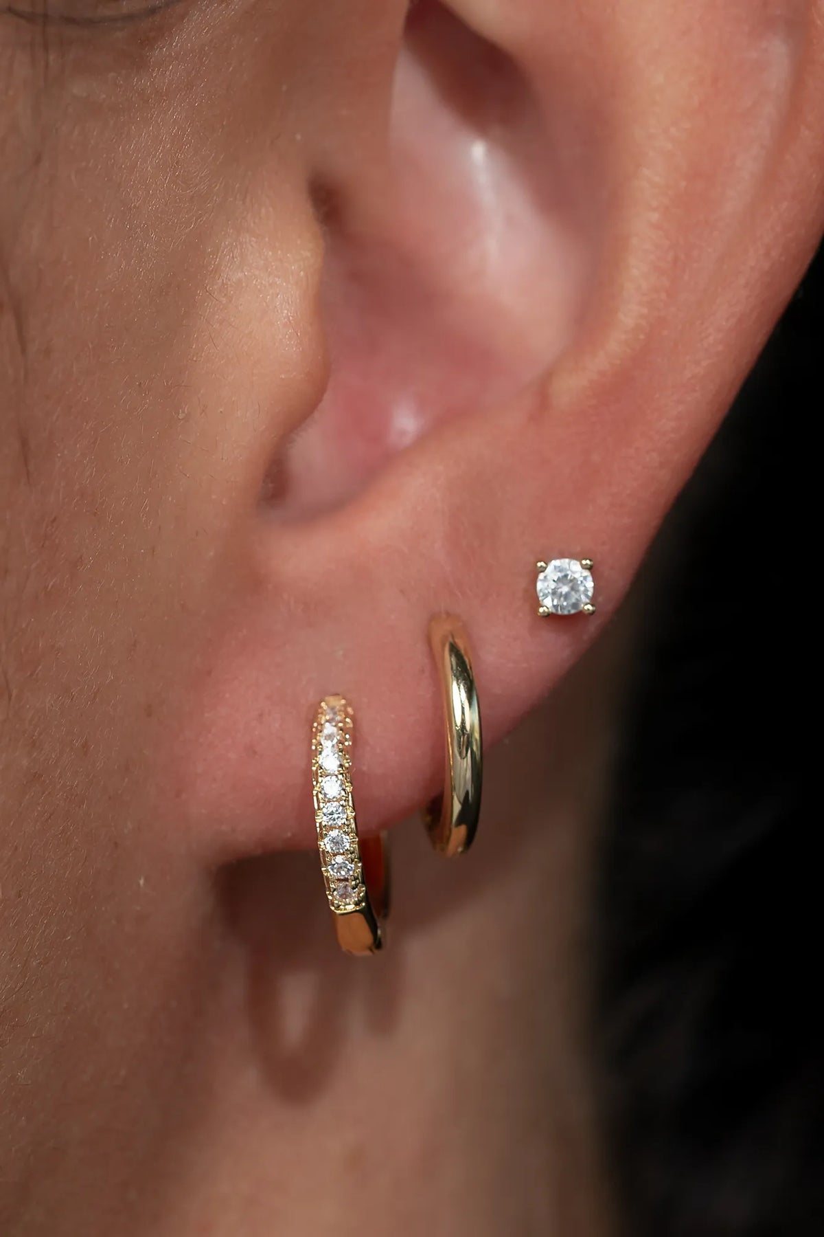 Model wearing a coordinated earring stack featuring gold-tone huggie hoop earrings with sparkling crystal accents, polished gold-tone huggie hoops, and round crystal stud earrings. 