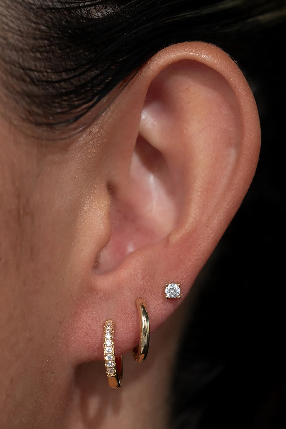 Model wearing a coordinated earring stack featuring gold-tone huggie hoop earrings with sparkling crystal accents, polished gold-tone huggie hoops, and round crystal stud earrings. 