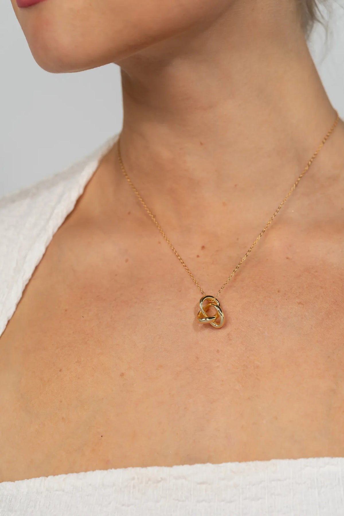Model wearing a delicate gold-tone necklace featuring an interlocking infinity knot pendant suspended from a fine chain. The minimalist design sits at the collarbone for an elegant, everyday look.
