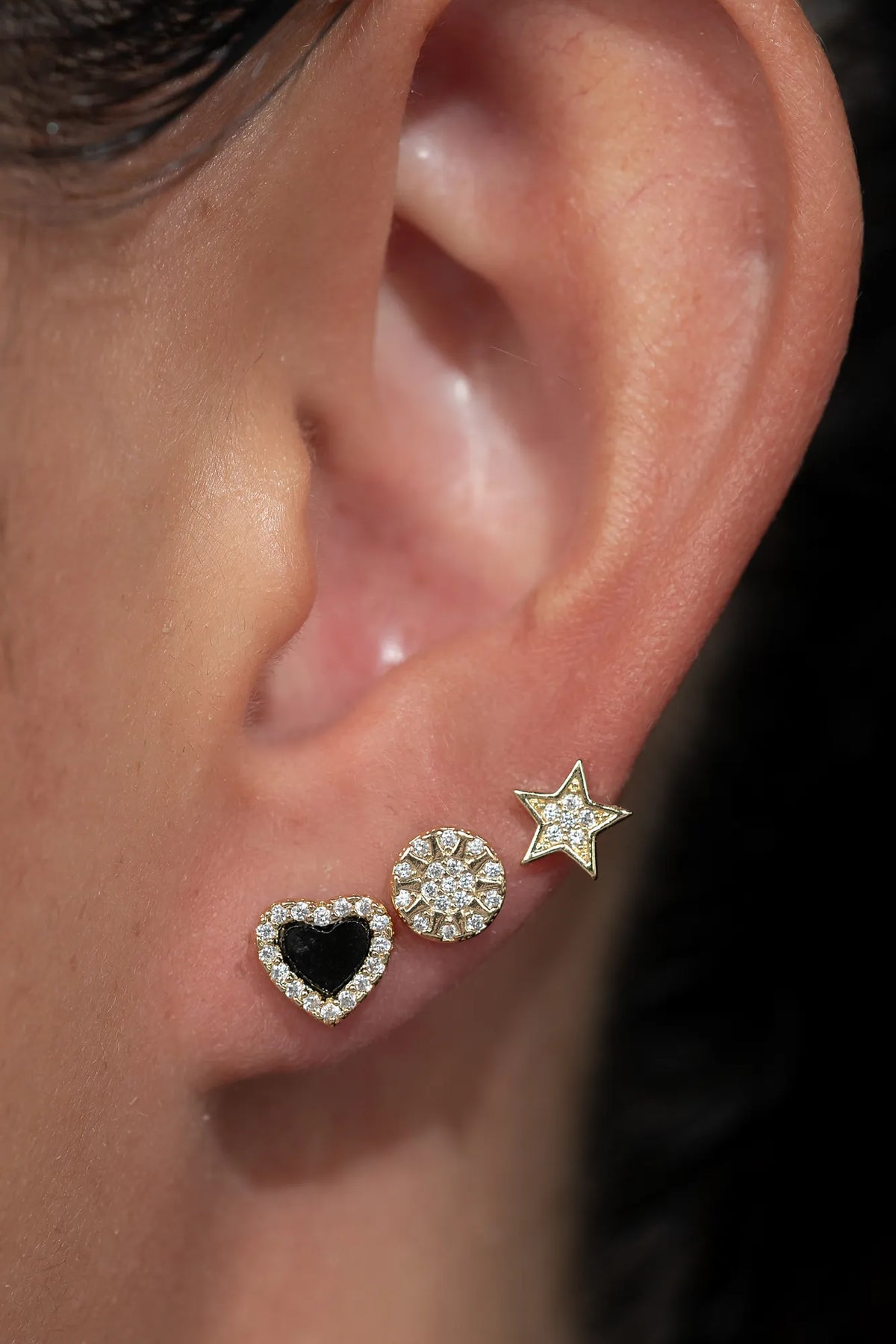 Model wearing three gold-tone stud earrings along the earlobe, including a black heart outlined with sparkling stones, a round sunburst-inspired crystal stud, and a star-shaped stud with crystal accents. The dainty celestial-inspired earrings create a layered, curated ear look with subtle sparkle and everyday versatility.