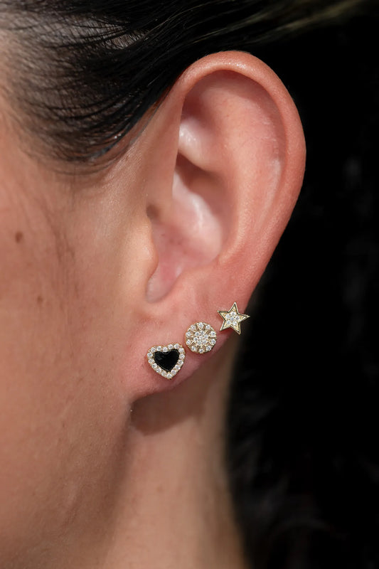 Model wearing three gold-tone stud earrings along the earlobe, including a black heart outlined with sparkling stones, a round sunburst-inspired crystal stud, and a star-shaped stud with crystal accents. The dainty celestial-inspired earrings create a layered, curated ear look with subtle sparkle and everyday versatility.