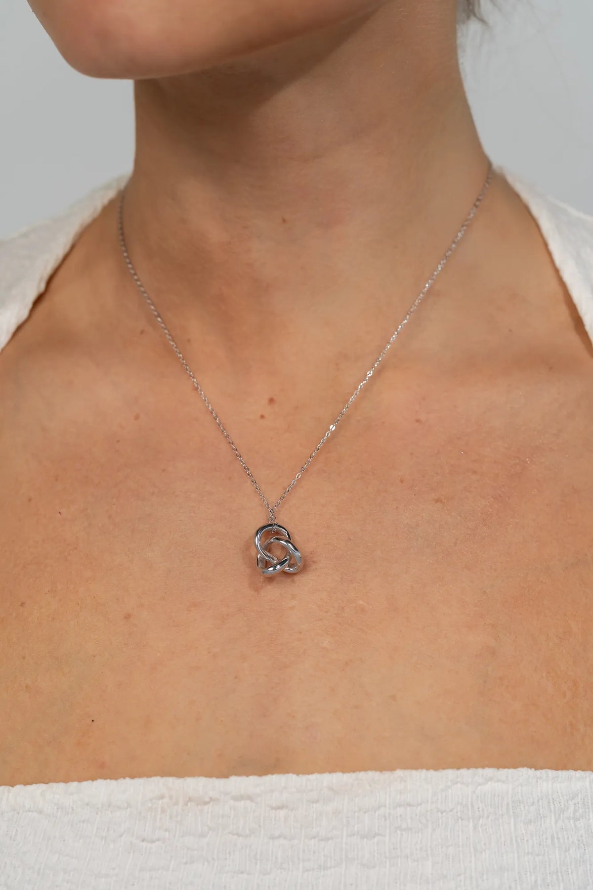 Model wearing a delicate silver-tone necklace featuring an interlocking infinity knot pendant suspended from a fine chain. The minimalist design sits at the collarbone for an elegant, everyday look.