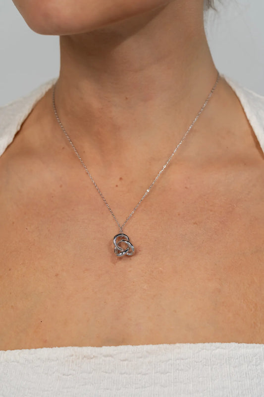 Model wearing a delicate silver-tone necklace featuring an interlocking infinity knot pendant suspended from a fine chain. The minimalist design sits at the collarbone for an elegant, everyday look.
