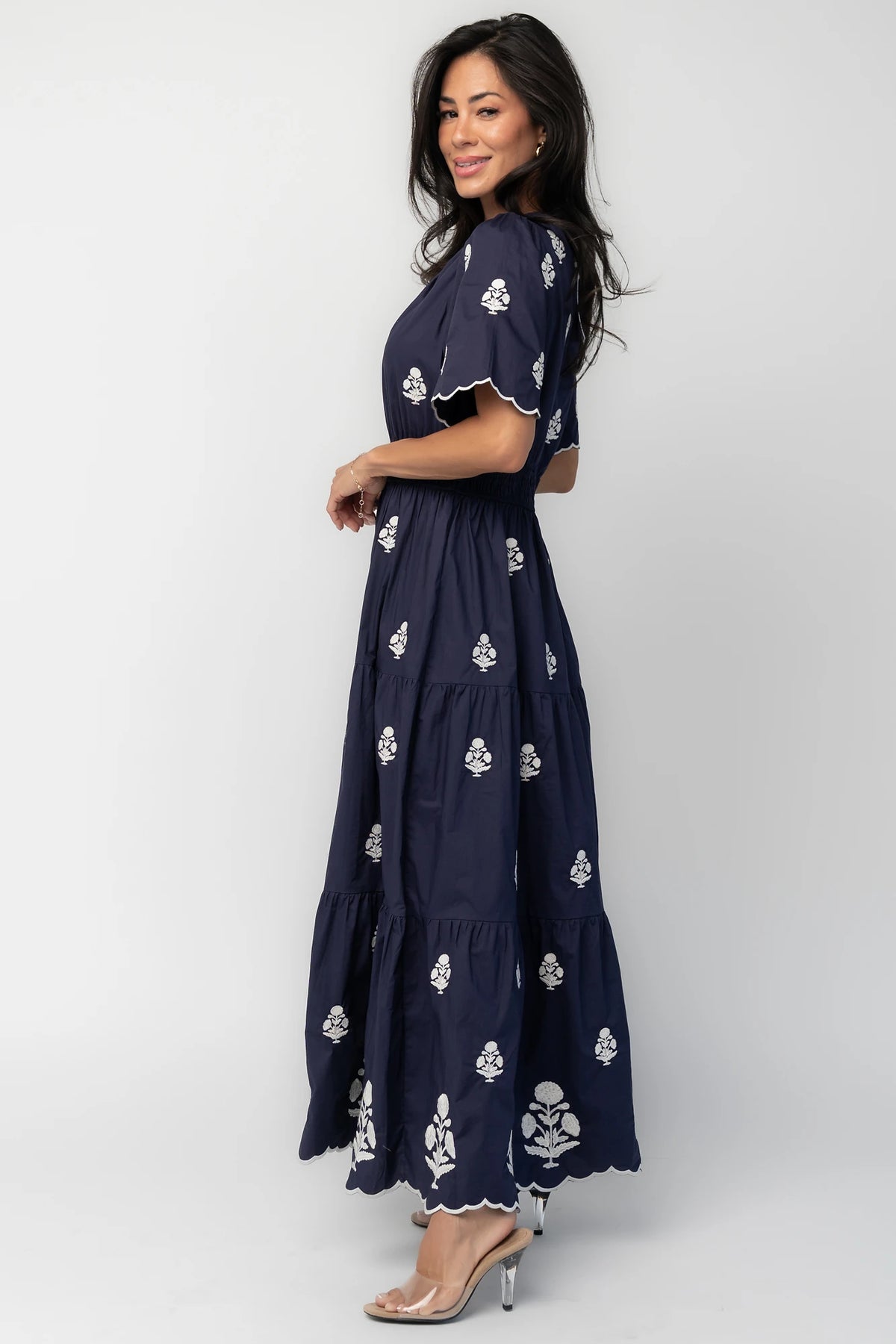 Model wearing the Delilah Embroidered Maxi Dress in navy featuring off-white floral embroidery, a split V-neckline, scalloped short sleeves, a smocked waistband, and a flowing tiered maxi skirt with a scalloped hem.