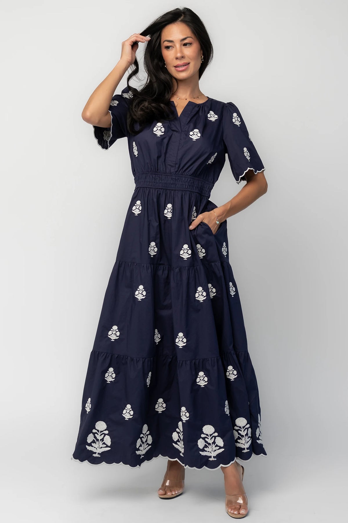 Model wearing the Delilah Embroidered Maxi Dress in navy featuring off-white floral embroidery, a split V-neckline, scalloped short sleeves, a smocked waistband, and a flowing tiered maxi skirt with a scalloped hem.