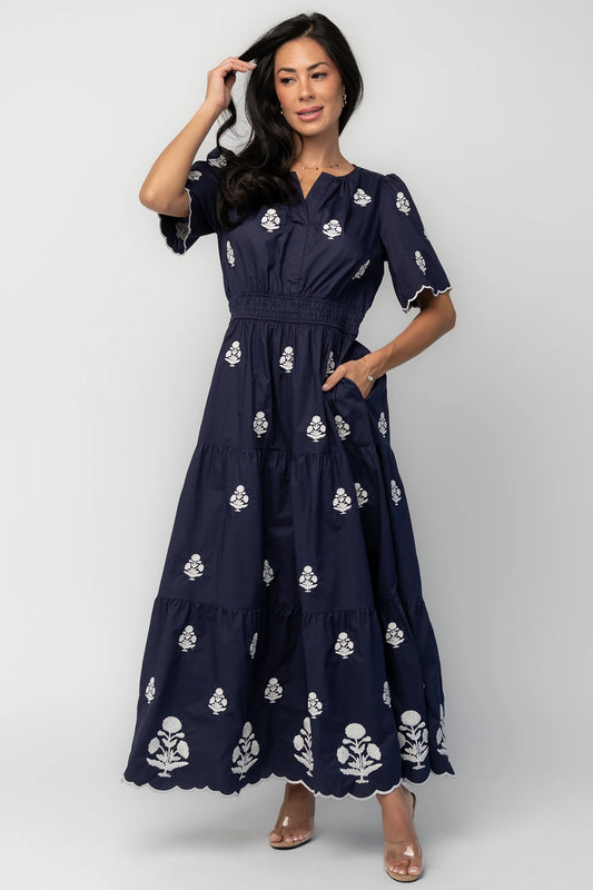 Model wearing the Delilah Embroidered Maxi Dress in navy featuring off-white floral embroidery, a split V-neckline, scalloped short sleeves, a smocked waistband, and a flowing tiered maxi skirt with a scalloped hem.