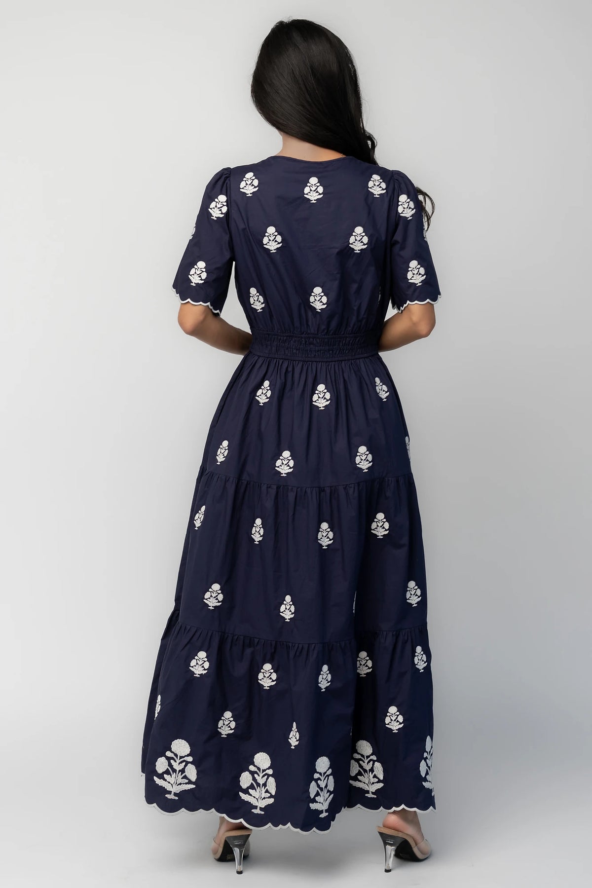Model wearing the Delilah Embroidered Maxi Dress in navy featuring off-white floral embroidery, a split V-neckline, scalloped short sleeves, a smocked waistband, and a flowing tiered maxi skirt with a scalloped hem.
