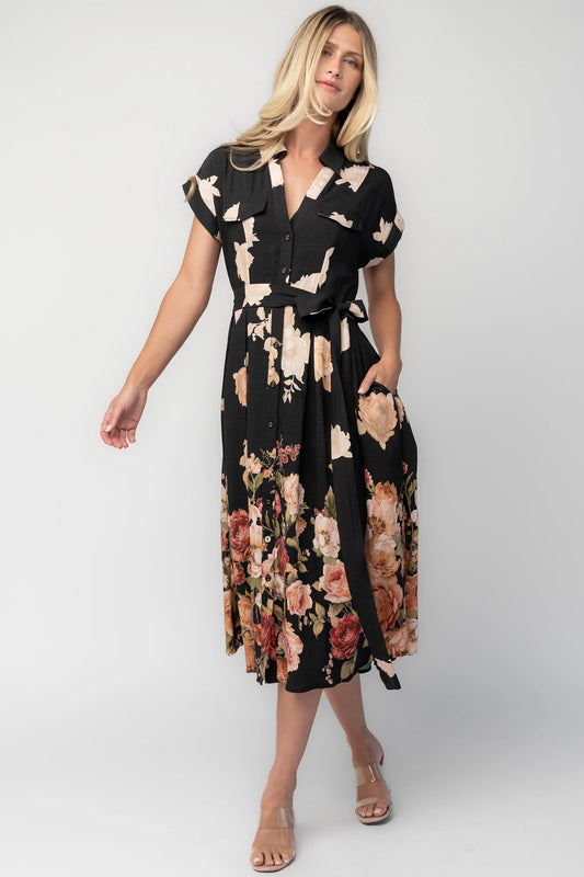 Model wearing a black floral midi dress with a collared neckline, functional button-front bodice, short sleeves, flap chest pockets, a removable waist tie, and a flowing midi-length skirt.