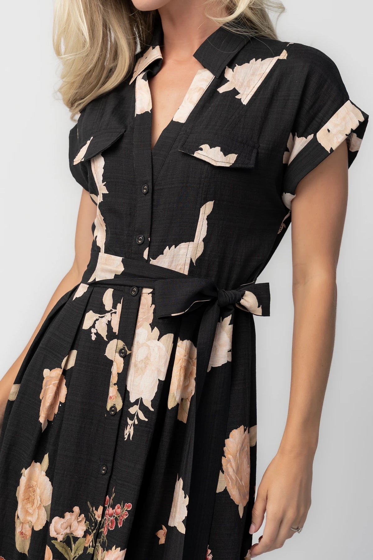Model wearing a black floral midi dress with a collared neckline, functional button-front bodice, short sleeves, flap chest pockets, a removable waist tie, and a flowing midi-length skirt.