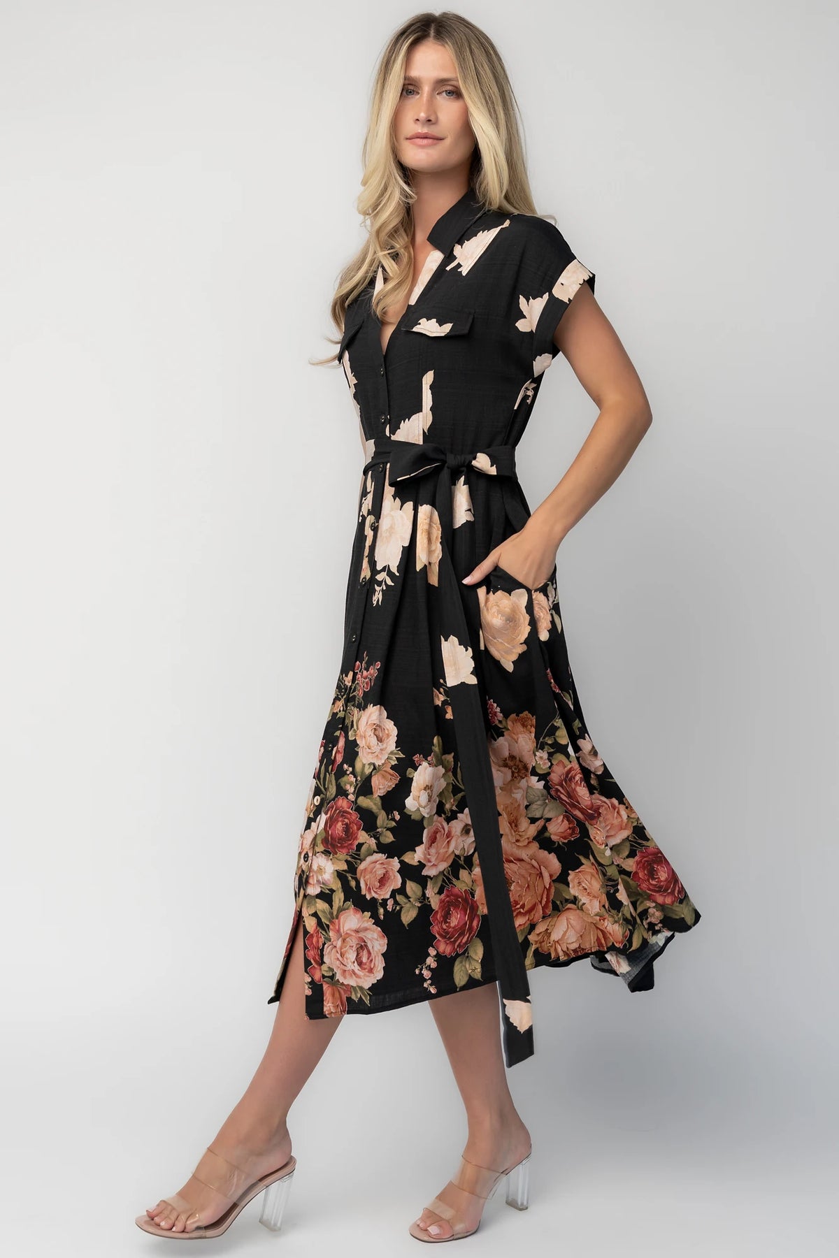 Model wearing a black floral midi dress with a collared neckline, functional button-front bodice, short sleeves, flap chest pockets, a removable waist tie, and a flowing midi-length skirt.