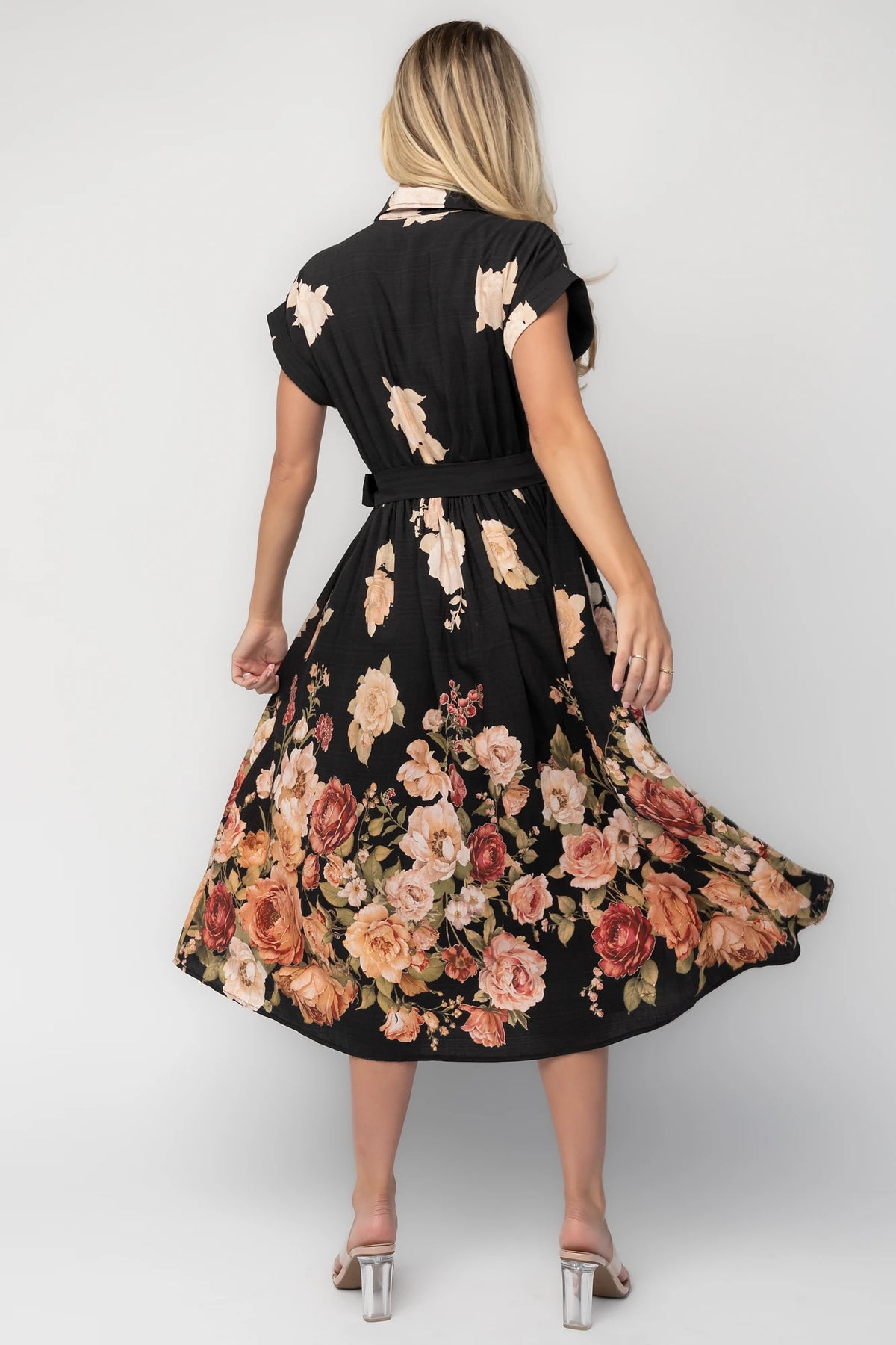 Model wearing a black floral midi dress with a collared neckline, functional button-front bodice, short sleeves, flap chest pockets, a removable waist tie, and a flowing midi-length skirt.