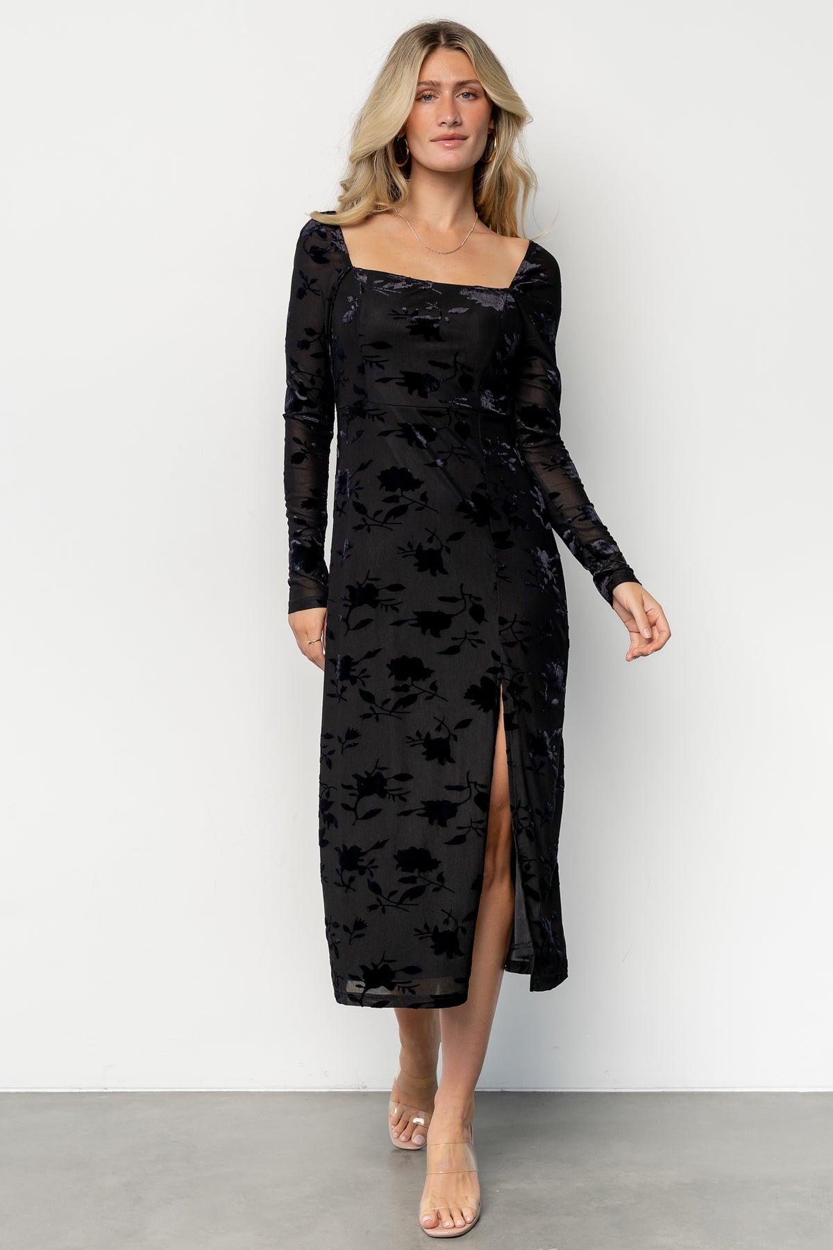 Front view of model wearing Tamara midi dress in black floral embossed velvet, square neckline, long sheer sleeves, and left leg slit.