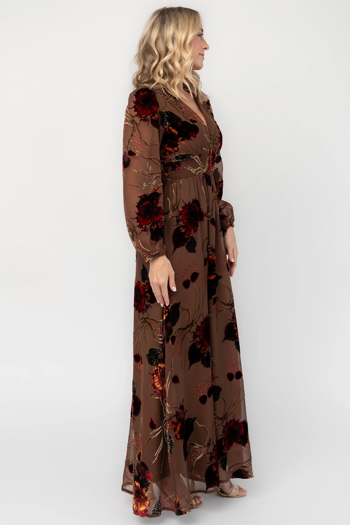 Brown burnout velvet maxi dress with a deep V-neckline, long sleeves, smocked waistband, and flowing asymmetrical hem with gold and burgundy floral accents.