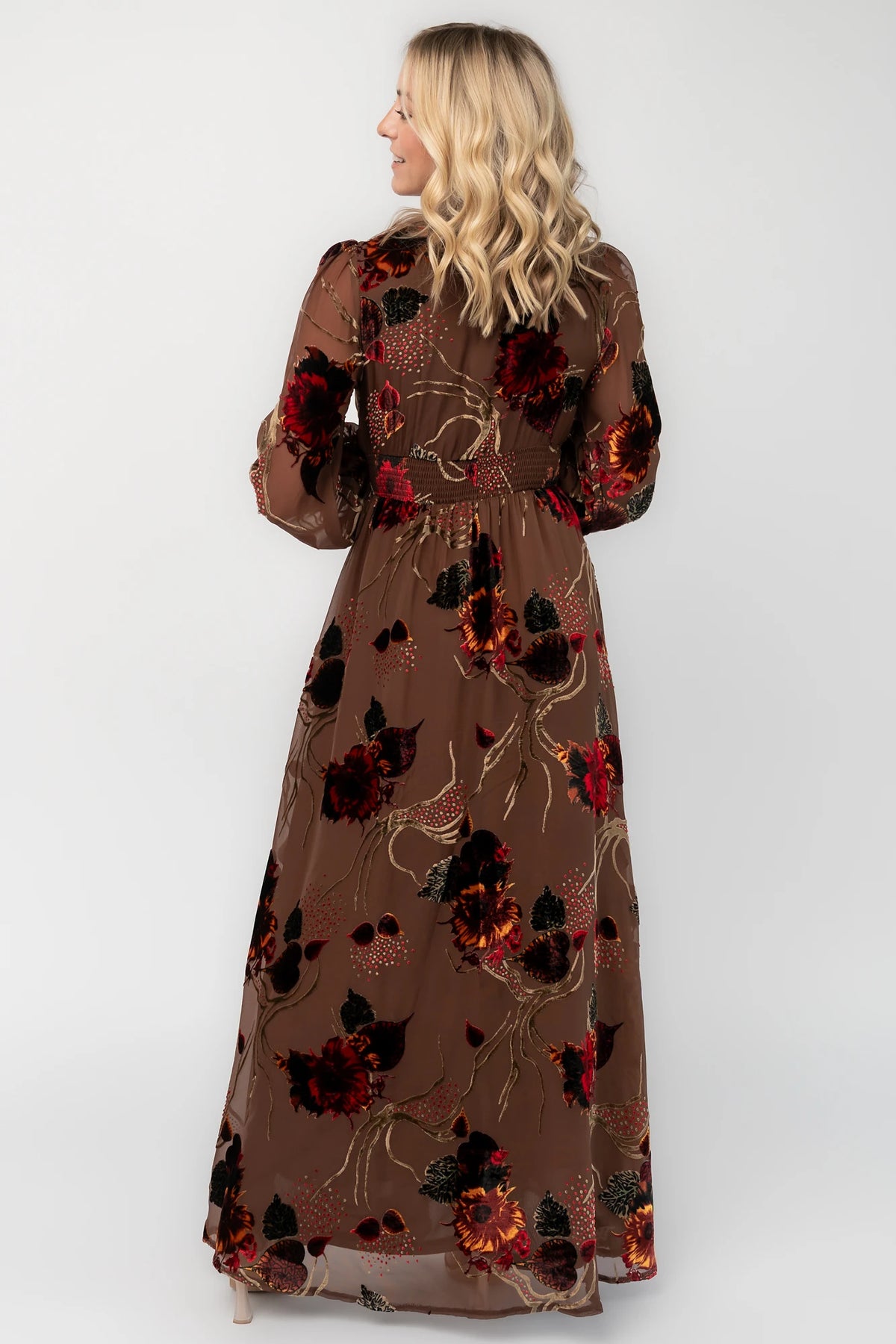Brown burnout velvet maxi dress with a deep V-neckline, long sleeves, smocked waistband, and flowing asymmetrical hem with gold and burgundy floral accents.