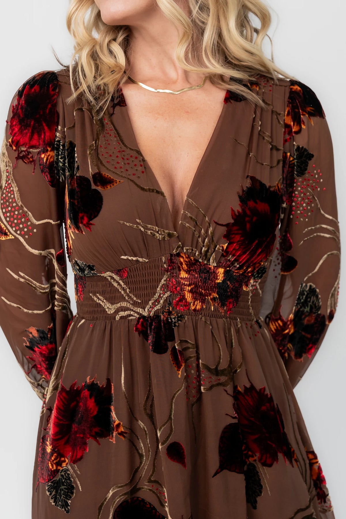 Brown burnout velvet maxi dress with a deep V-neckline, long sleeves, smocked waistband, and flowing asymmetrical hem with gold and burgundy floral accents.
