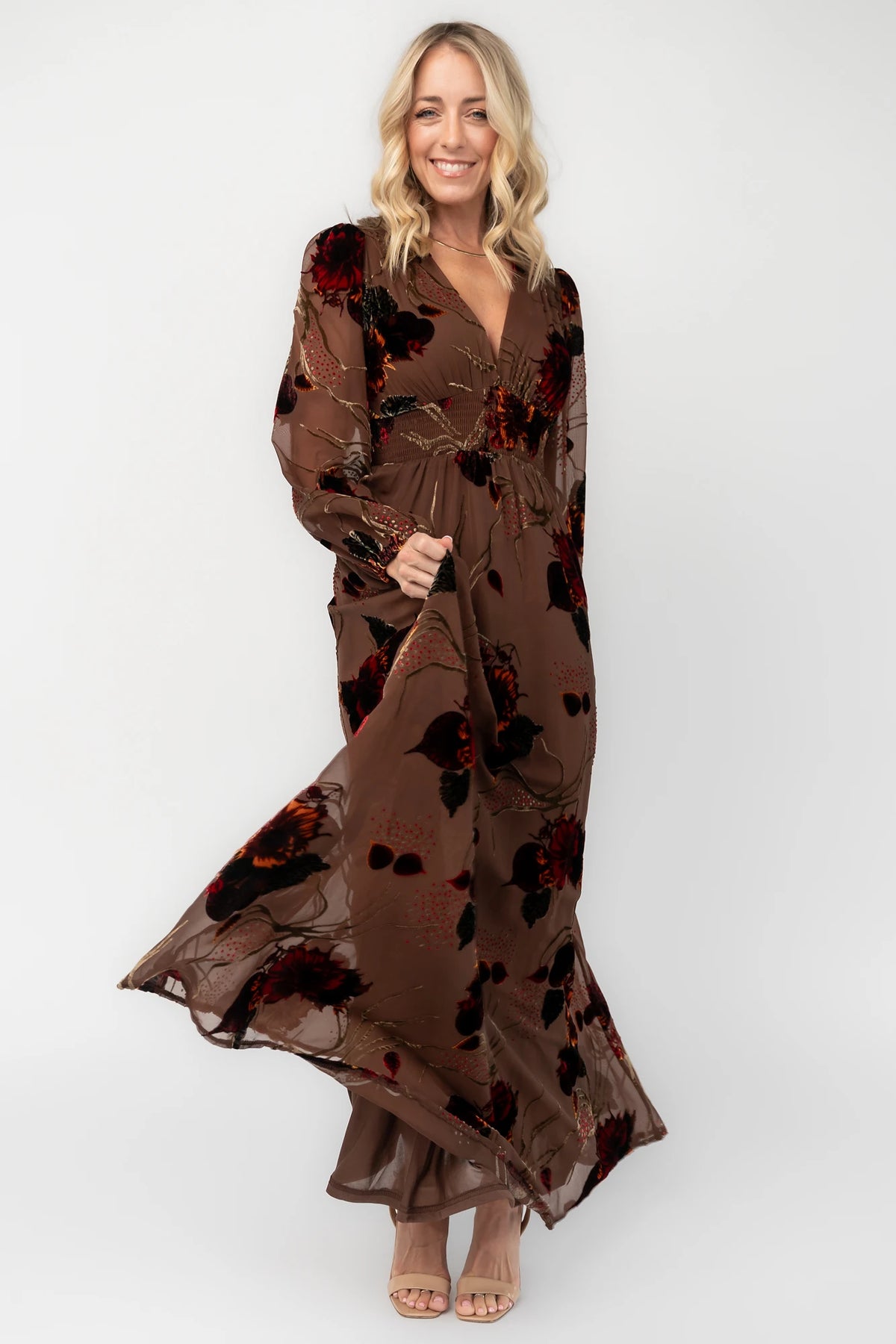Brown burnout velvet maxi dress with a deep V-neckline, long sleeves, smocked waistband, and flowing asymmetrical hem with gold and burgundy floral accents.